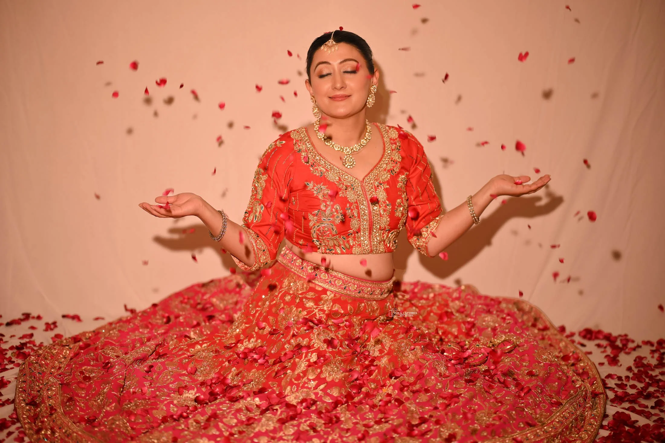 A woman in a red and gold traditional dress is seated cross-legged, surrounded by falling rose petals, with her eyes closed and a serene expression.