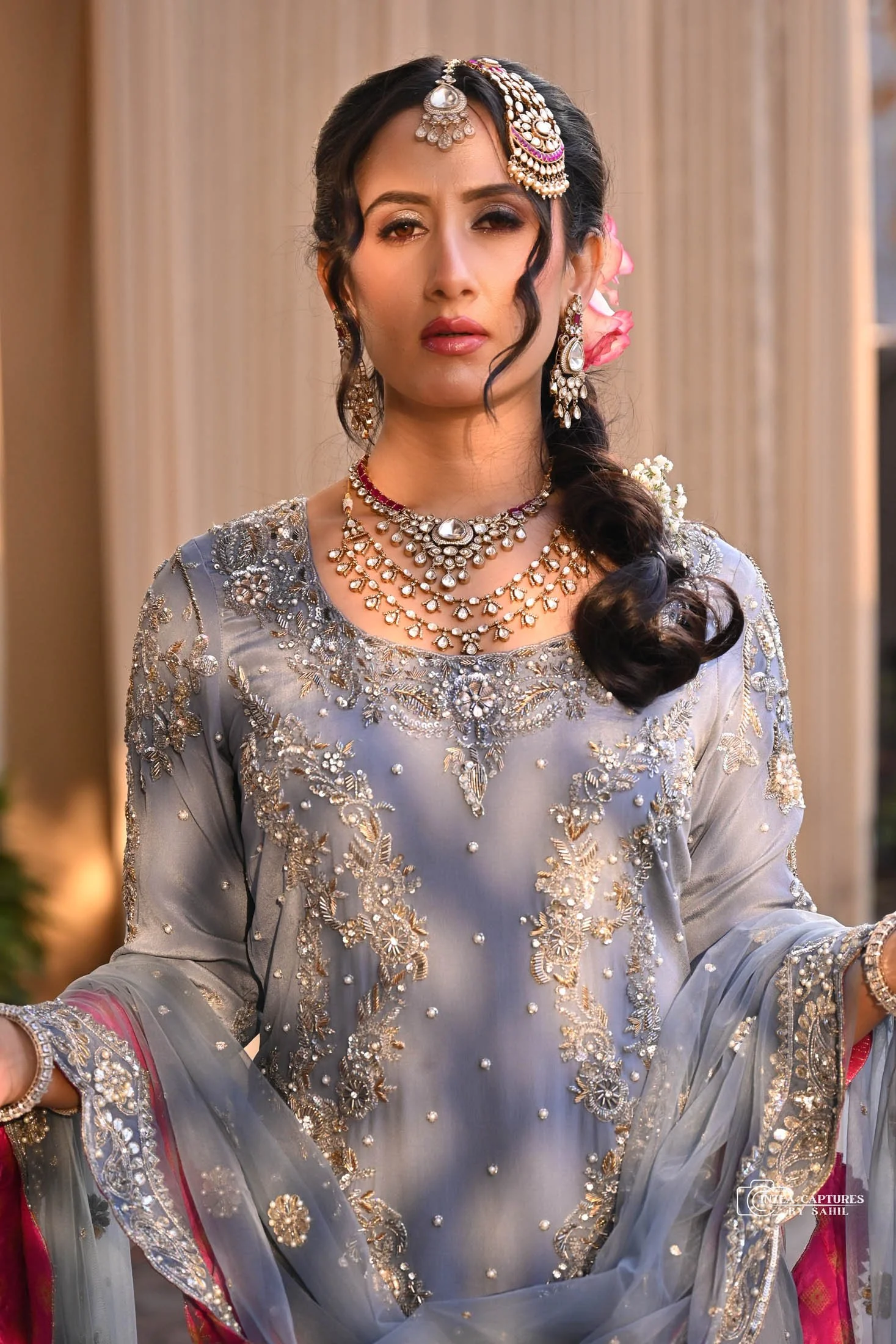 A woman dressed in a traditional, heavily embellished grey outfit with intricate embroidery and pearls, wearing elaborate jewelry including necklaces, earrings, and a headpiece, standing indoors with a neutral background.