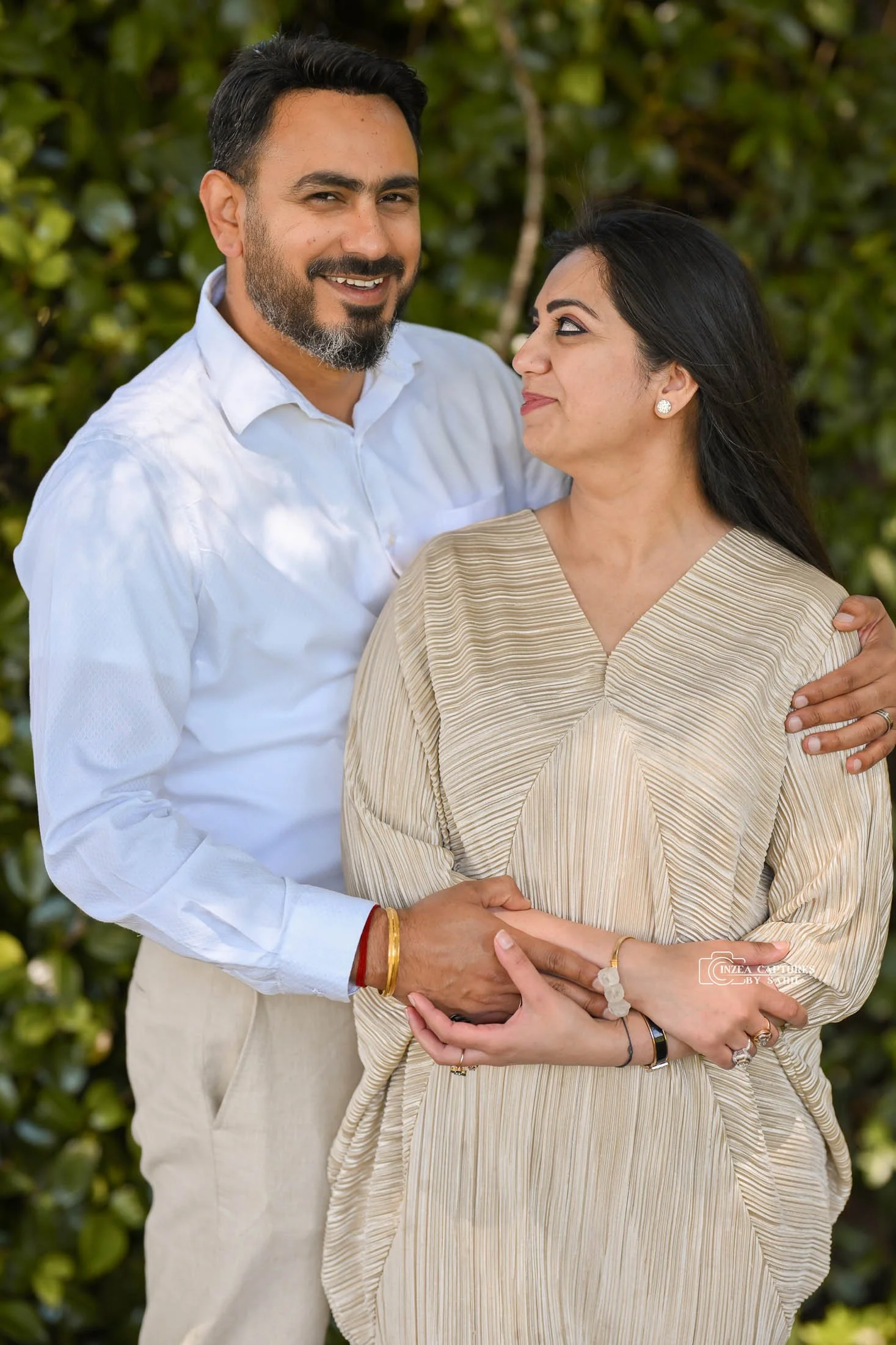 A man and woman standing close together outdoors, smiling and looking at each other. The man has dark hair and a beard, wearing a white shirt. The woman has long dark hair, wearing a beige striped dress with jewelry.