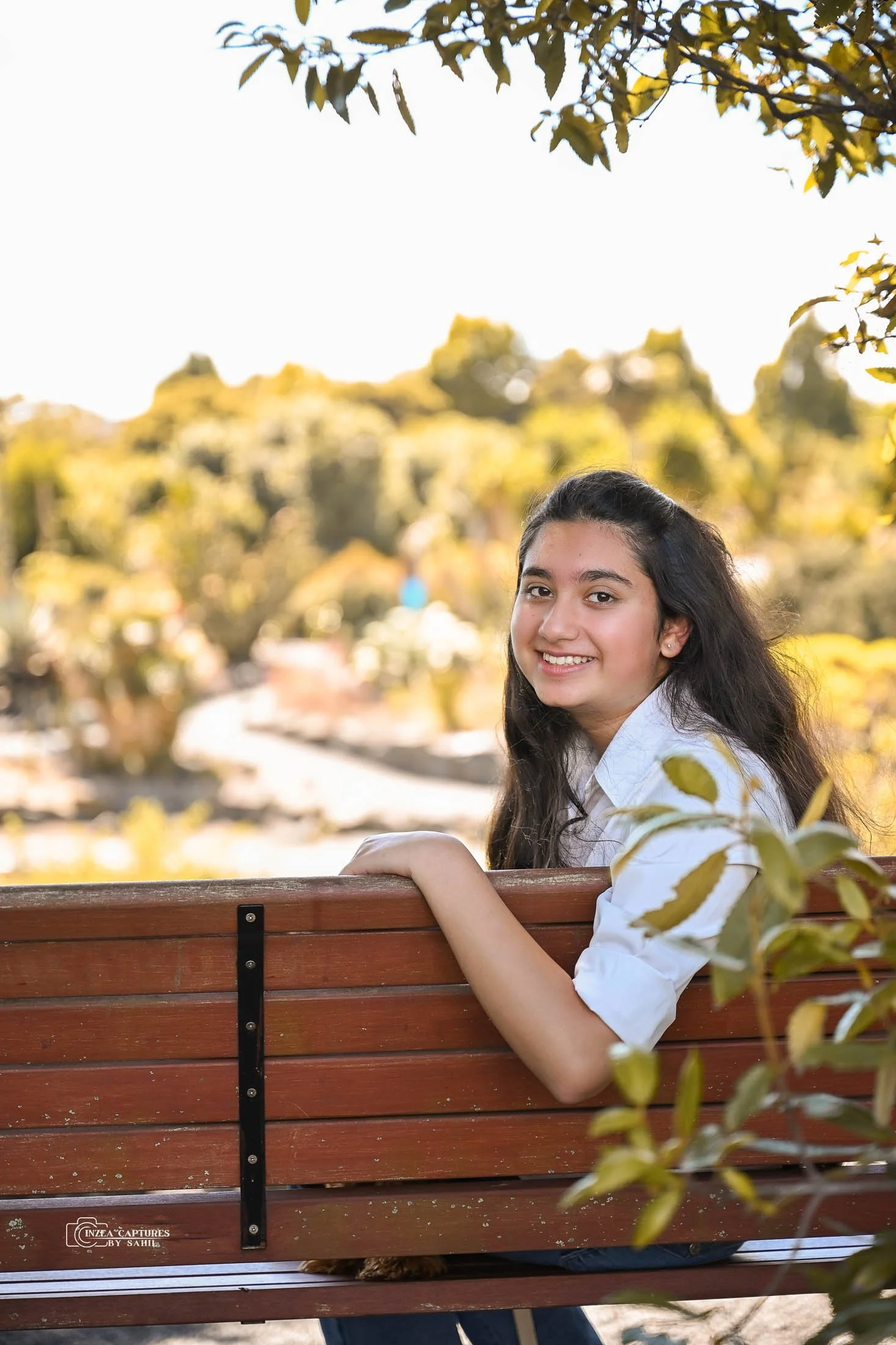 A young girl with long black hair, wearing a white shirt, sitting on a wooden park bench, smiling at the camera with trees and a bright sky in the background.