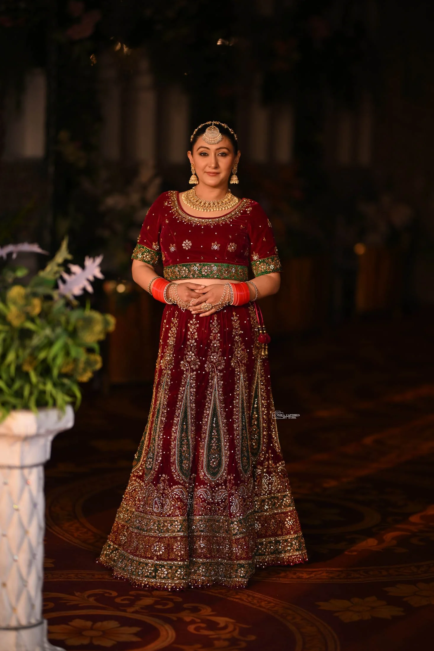 A woman dressed in a traditional ornate red and green Indian bridal lehenga with intricate embroidery, standing indoors with dim lighting and a floral arrangement partially visible on the left.