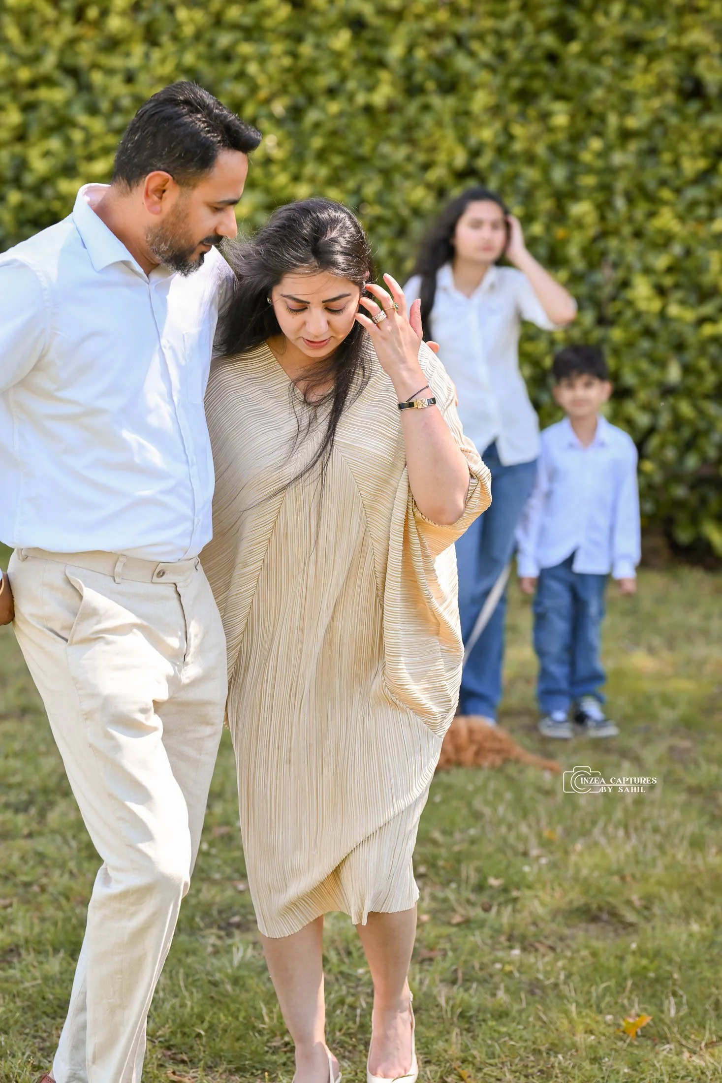 A middle-aged woman in a beige dress with long dark hair is being comforted by a man in a white shirt and beige pants outdoors. In the background, a woman and two children are standing near a hedge, with the woman holding her head and the children lo