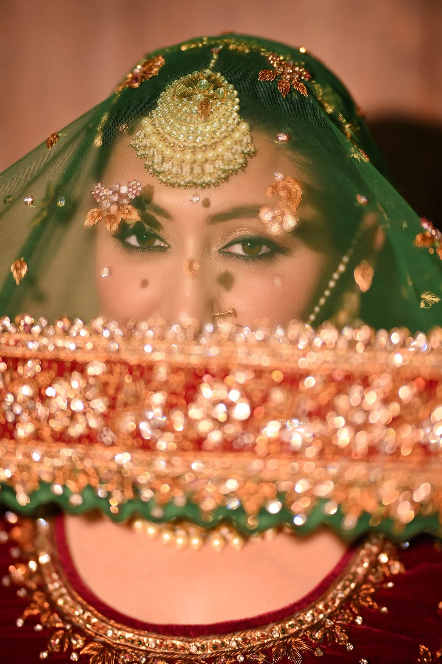 A woman wearing traditional Indian bridal attire with intricate jewelry, a green veil, and a maroon dress, partially obscured by a decorative, bejeweled headpiece.