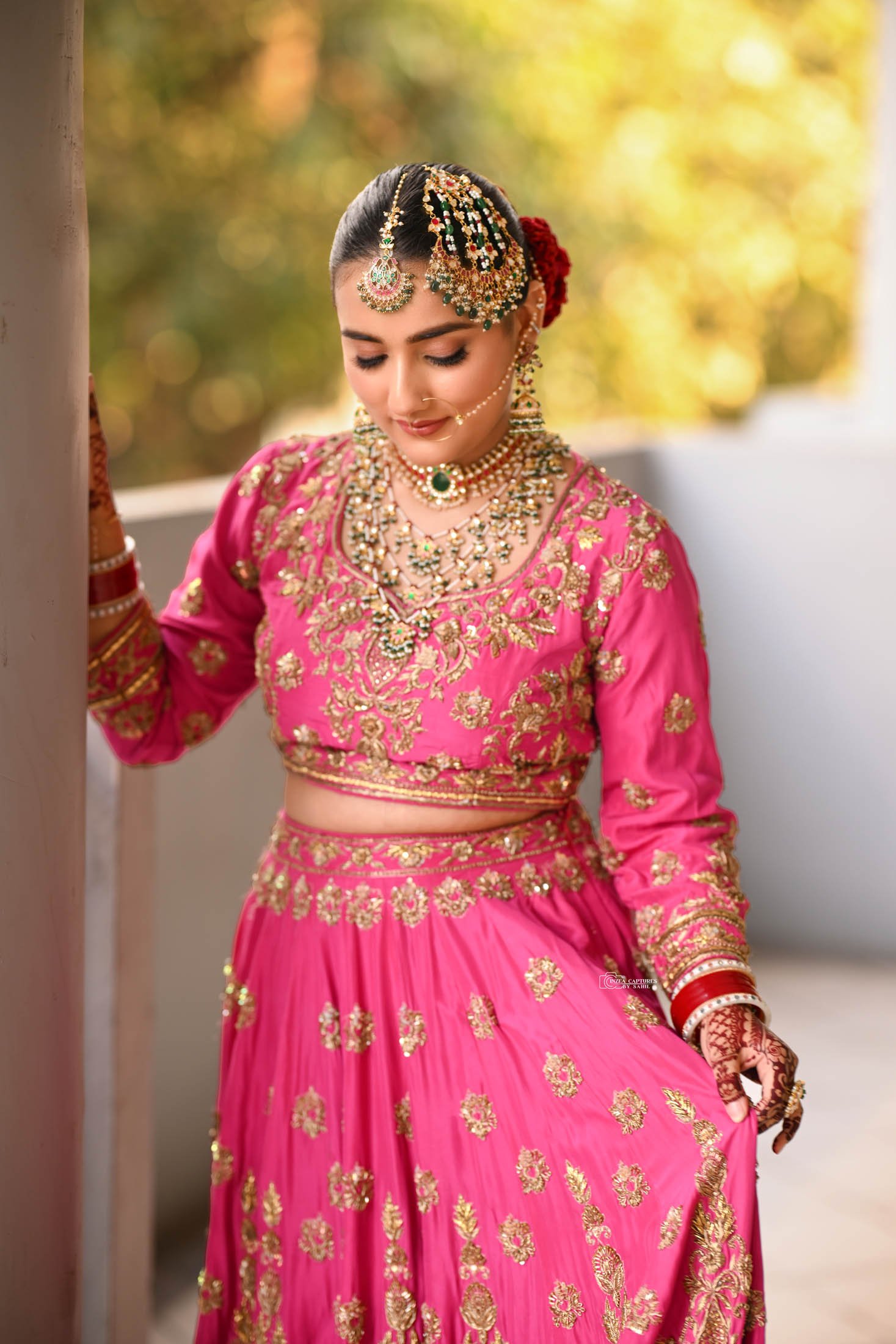 A woman dressed in traditional Indian pink bridal attire, adorned with gold embroidery, and wearing elaborate jewelry including a necklace, earrings, nose ring, and accessories in her hair and arms.