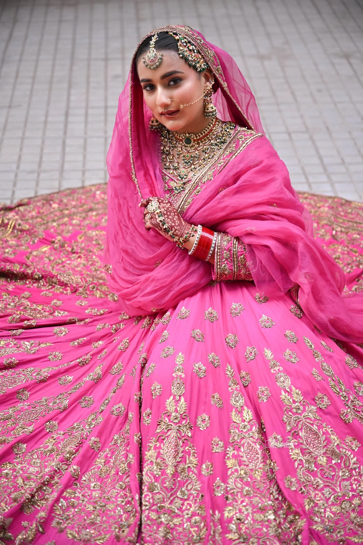 A woman dressed in a traditional pink and gold Indian bridal outfit, sitting on the ground, wearing elaborate jewelry, including a headpiece, earrings, necklace, nose ring, bangles, and henna on her hands, with a tiled floor in the background.