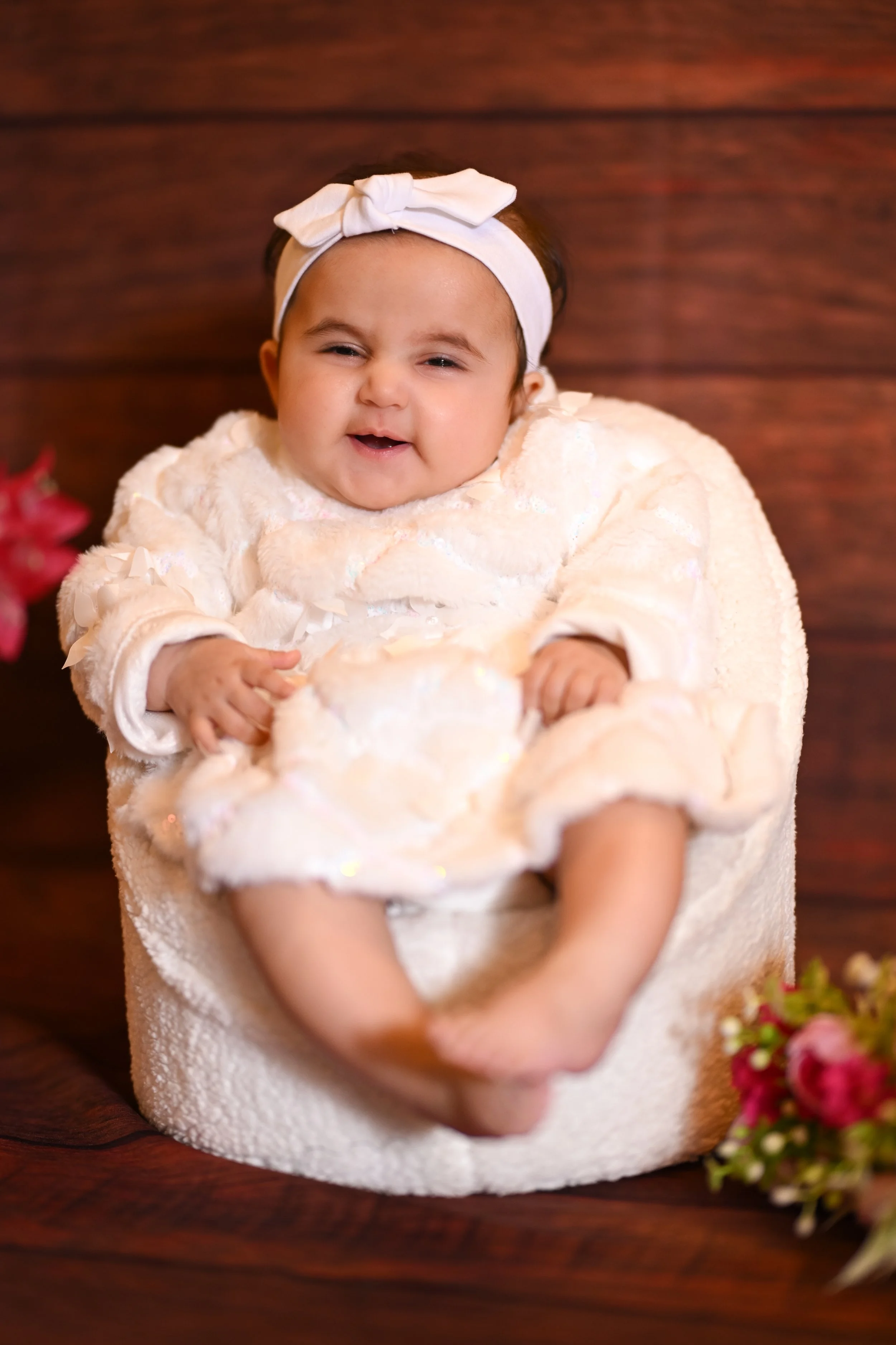 Smiling baby girl sitting in a white plush chair, wearing a white headband with a bow, dressed in a cream-colored outfit, with flowers nearby on a wooden surface.