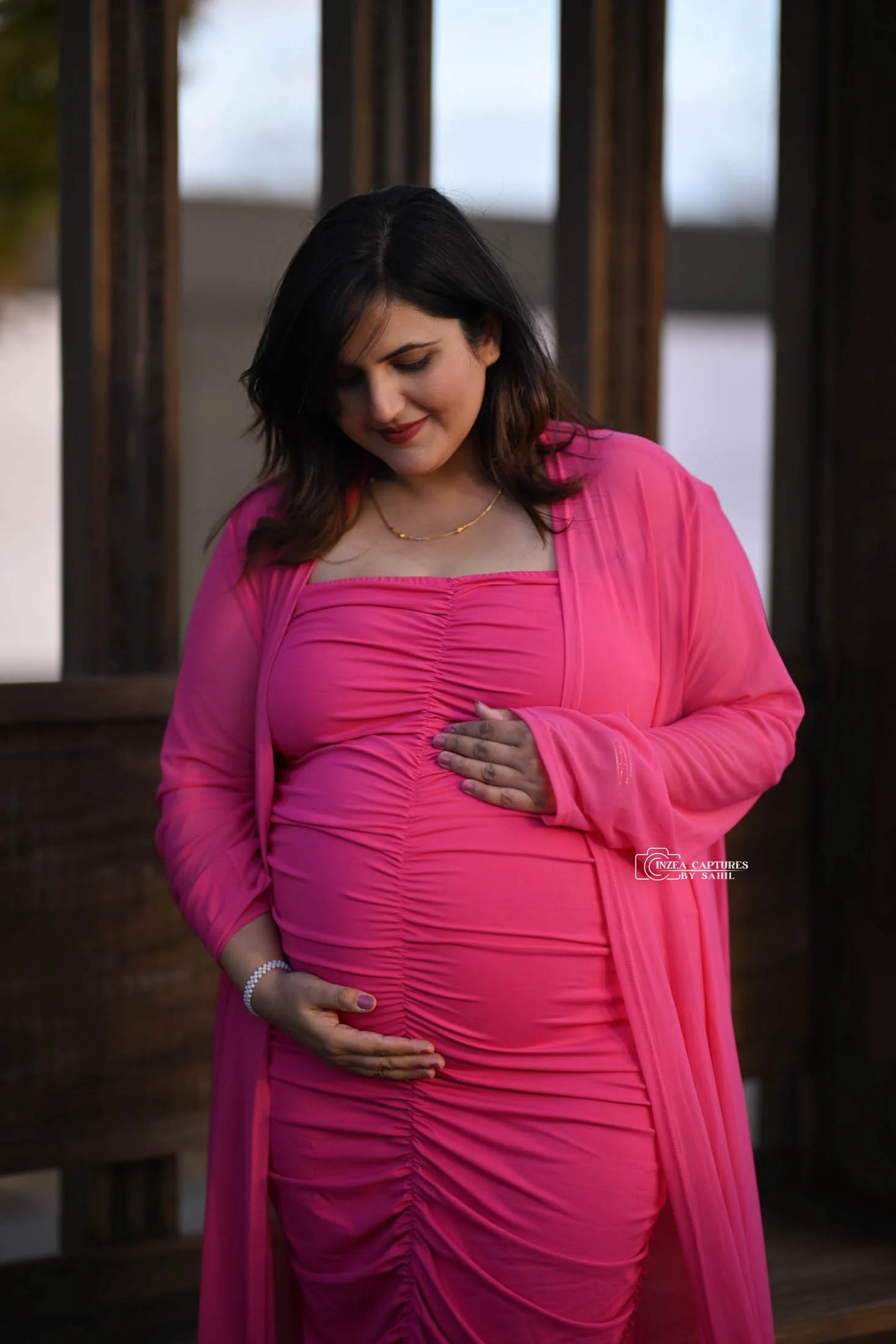 Pregnant woman in a pink dress touching her belly and looking down, standing outdoors.