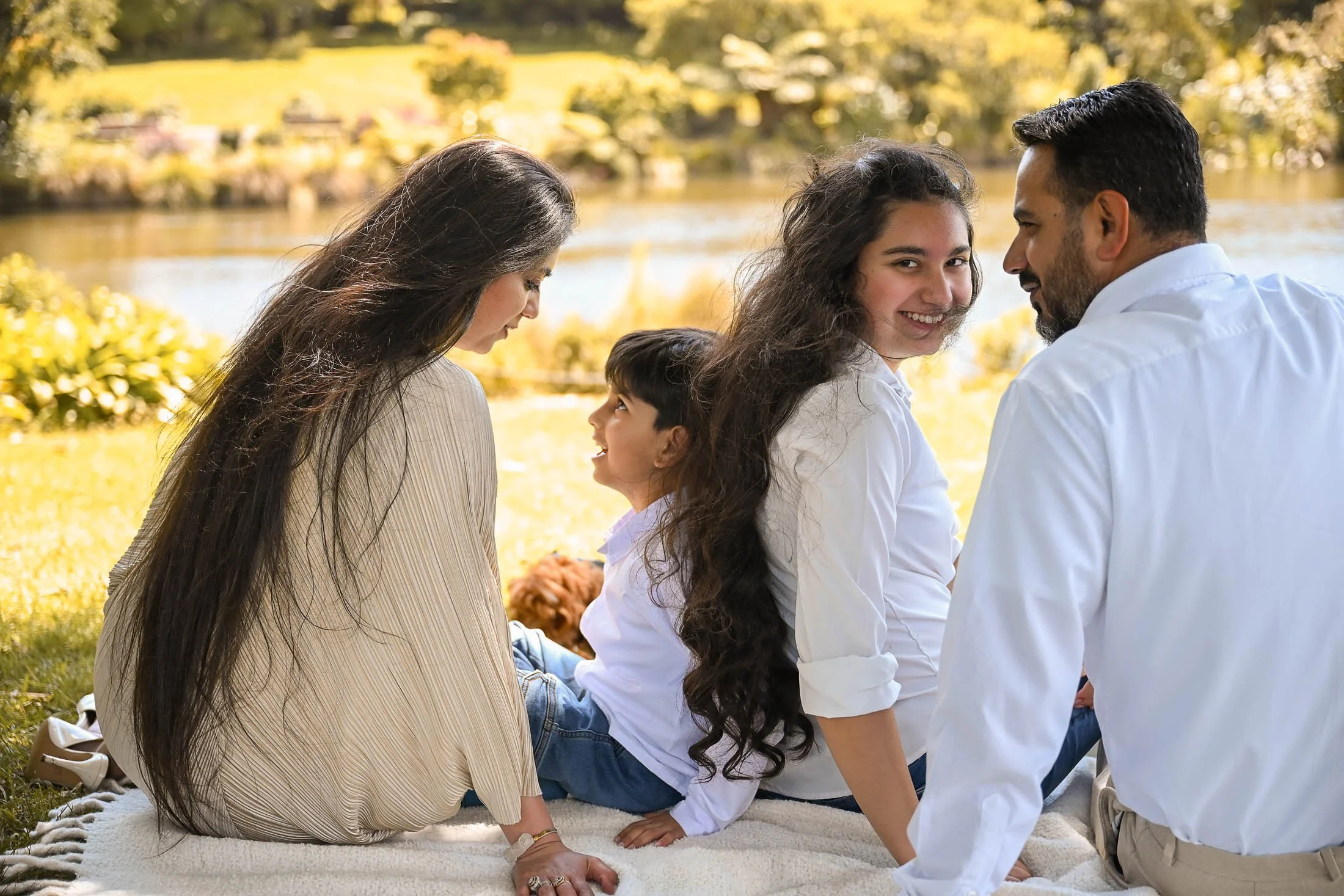 A family of four with a young girl, woman, man, and an older girl sitting outdoors on a blanket near a lake with trees and yellow foliage in the background.