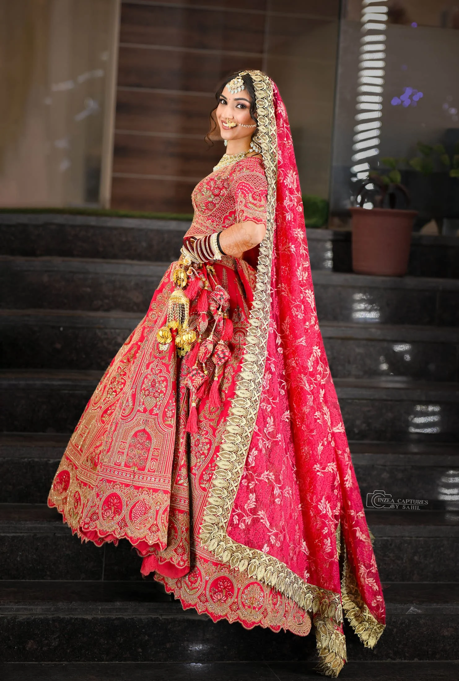 A woman in a traditional red and gold Indian bridal outfit, smiling, standing on dark stone steps with potted plants and a wooden wall in the background.