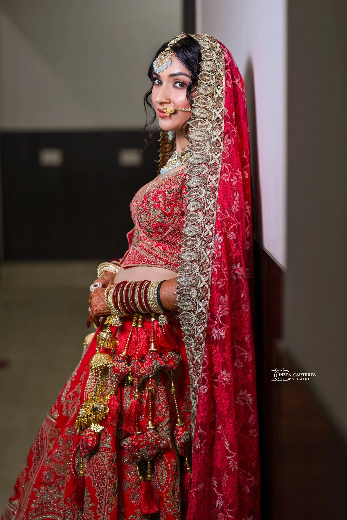 A woman dressed in traditional Indian bridal attire, wearing a red heavily embroidered lehenga with gold accents, a matching dupatta with intricate silver embroidery, and adorned with jewelry including bangles, necklace, nose ring, and headpiece, sta