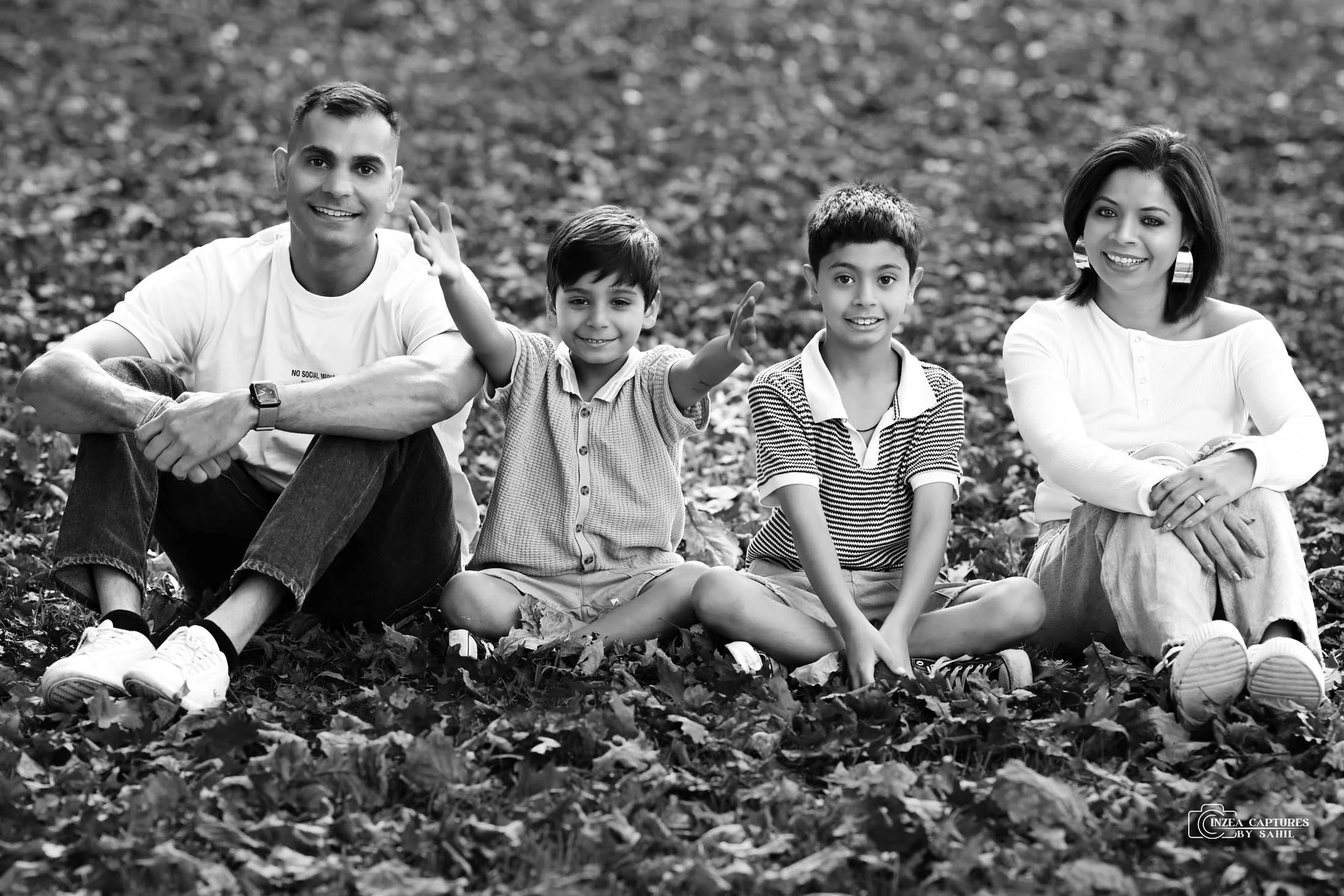 A family of four sitting on the ground covered with fallen leaves outdoors, smiling and waving at the camera in a black-and-white photo.