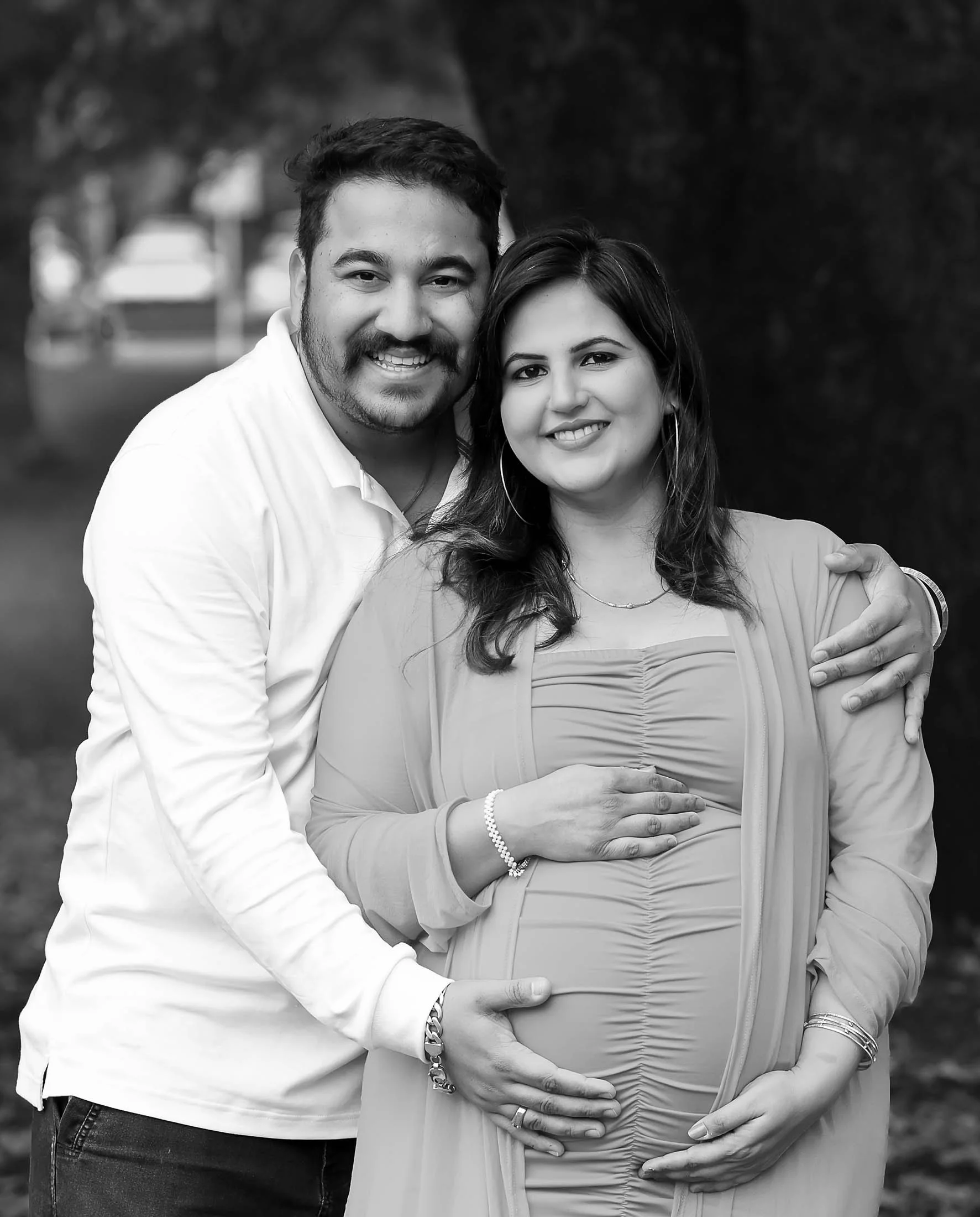 A happy couple, with the man hugging the pregnant woman from behind, standing outdoors with trees in the background, smiling at the camera.