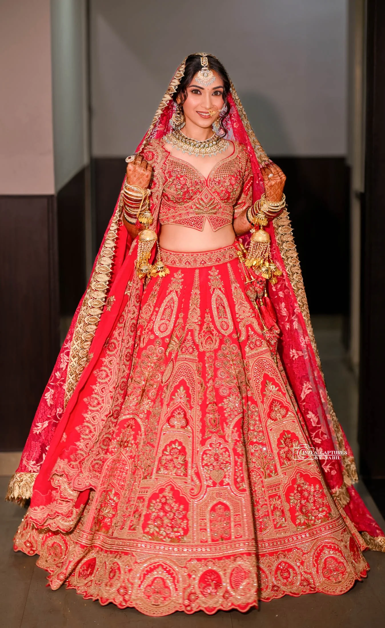 A woman dressed in a traditional red and gold bridal Indian outfit, including a heavily embroidered lehenga, choli, and dupatta, adorned with gold jewelry and bangles, standing indoors.