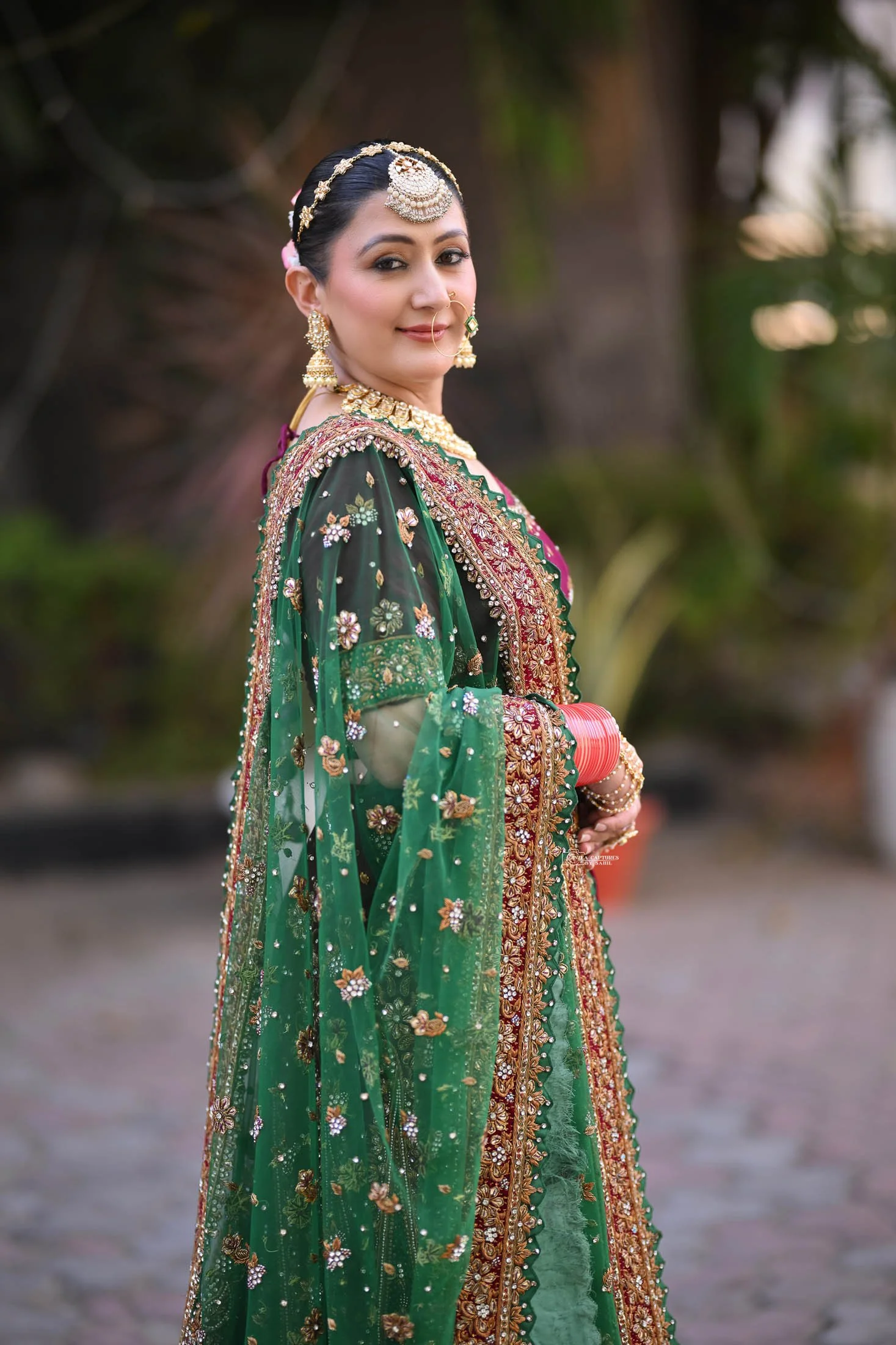 A woman dressed in a traditional Indian green embroidered saree with gold and gemstone jewelry, standing outdoors with blurred foliage in the background.