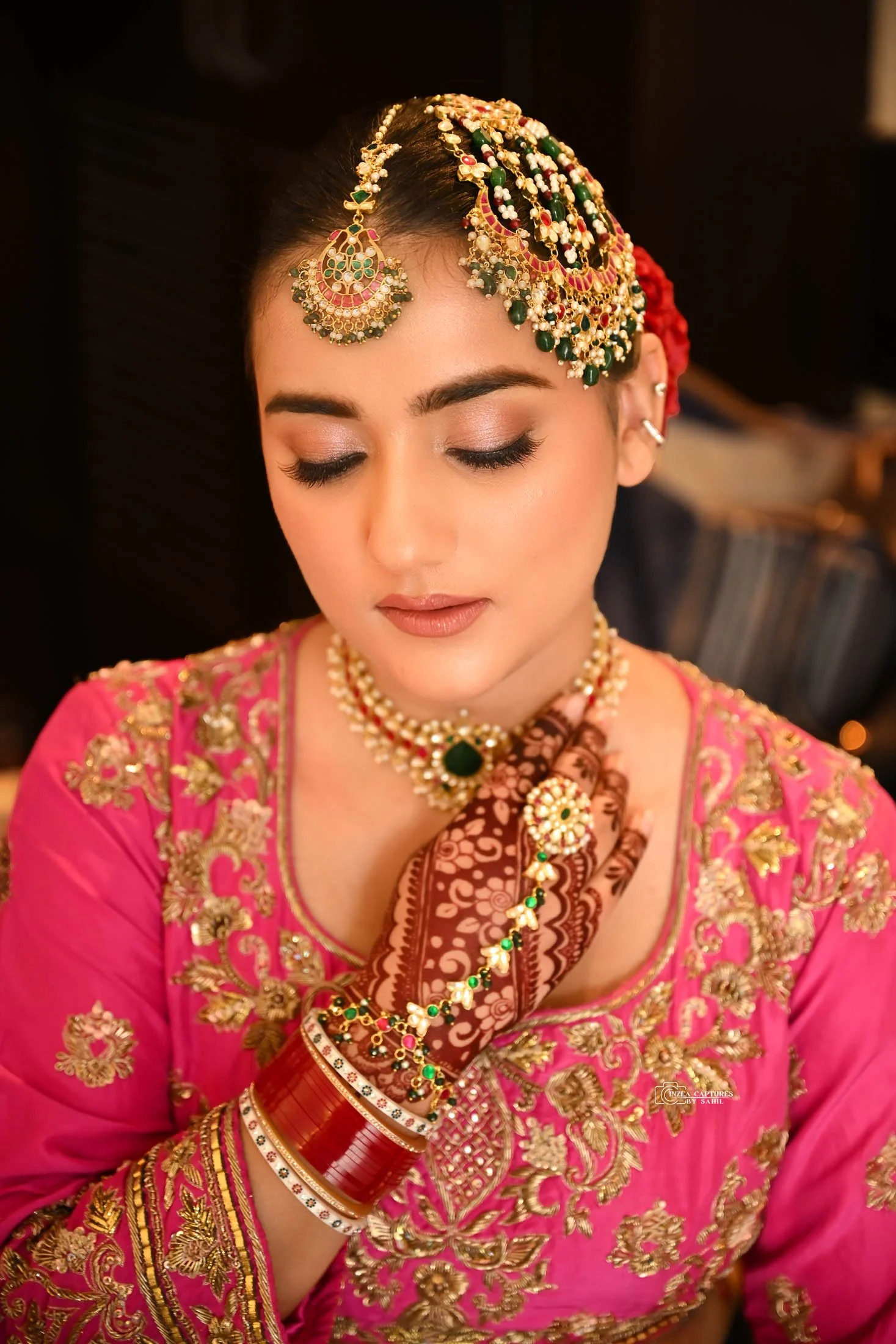 Woman in traditional Indian bridal attire with gold jewelry, pink embroidered dress, and henna on her hand, with her eyes closed.