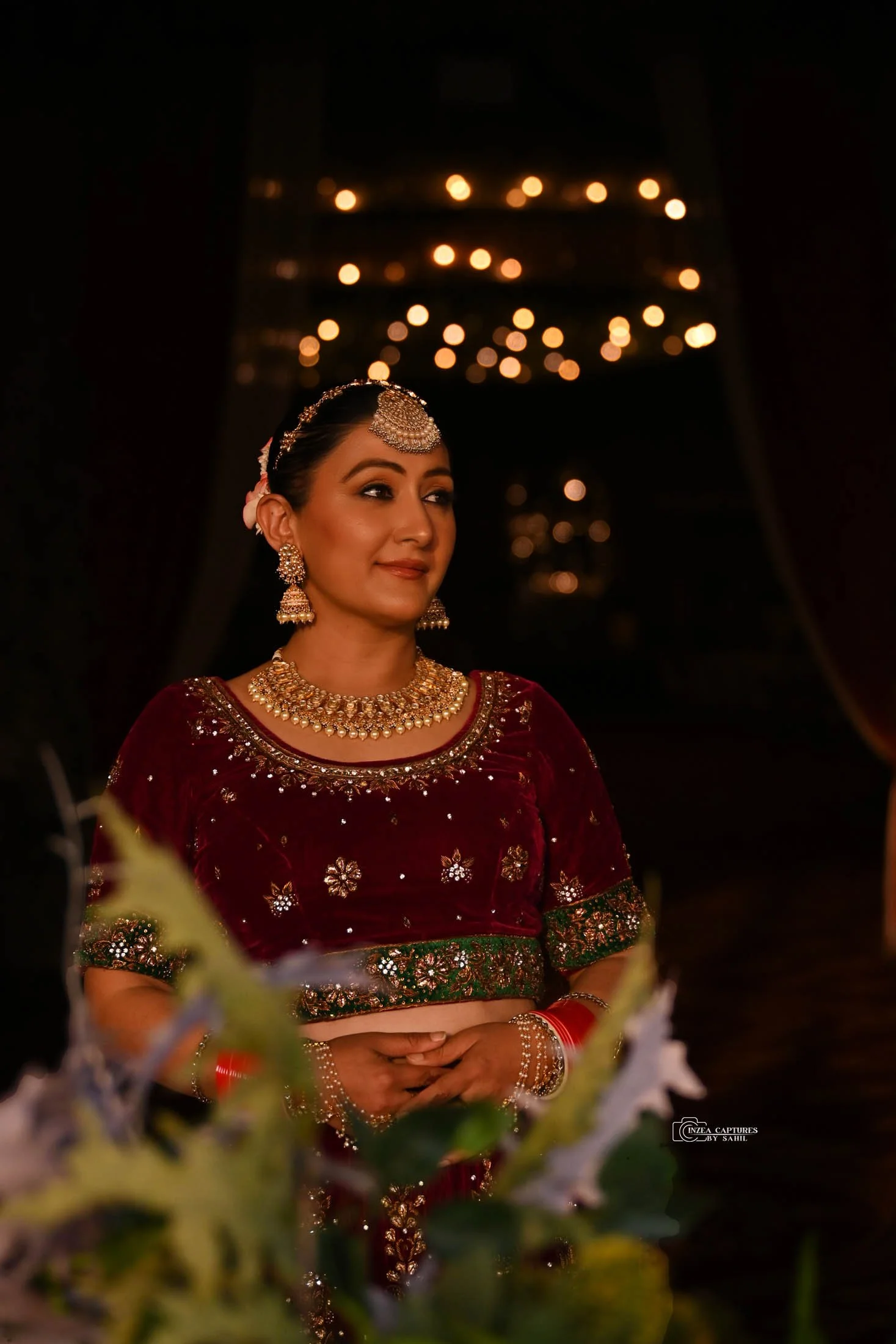 A woman dressed in traditional Indian attire, wearing ornate jewelry, standing near flowers with a dark background and warm lighting.