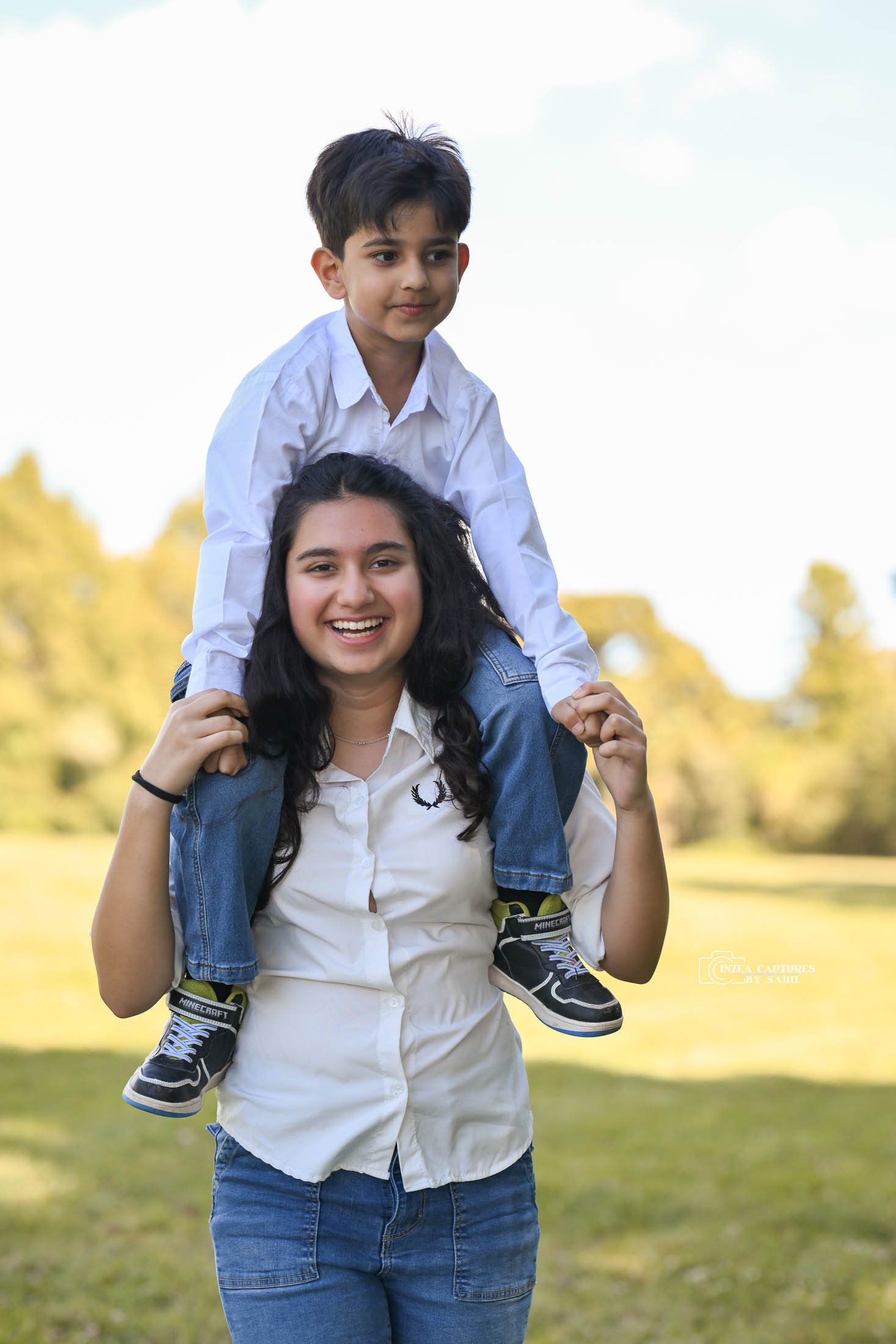 A young woman with dark curly hair smiling, giving a piggyback ride to a young boy with short dark hair on her shoulders, outdoors in a park with trees and grass in the background.
