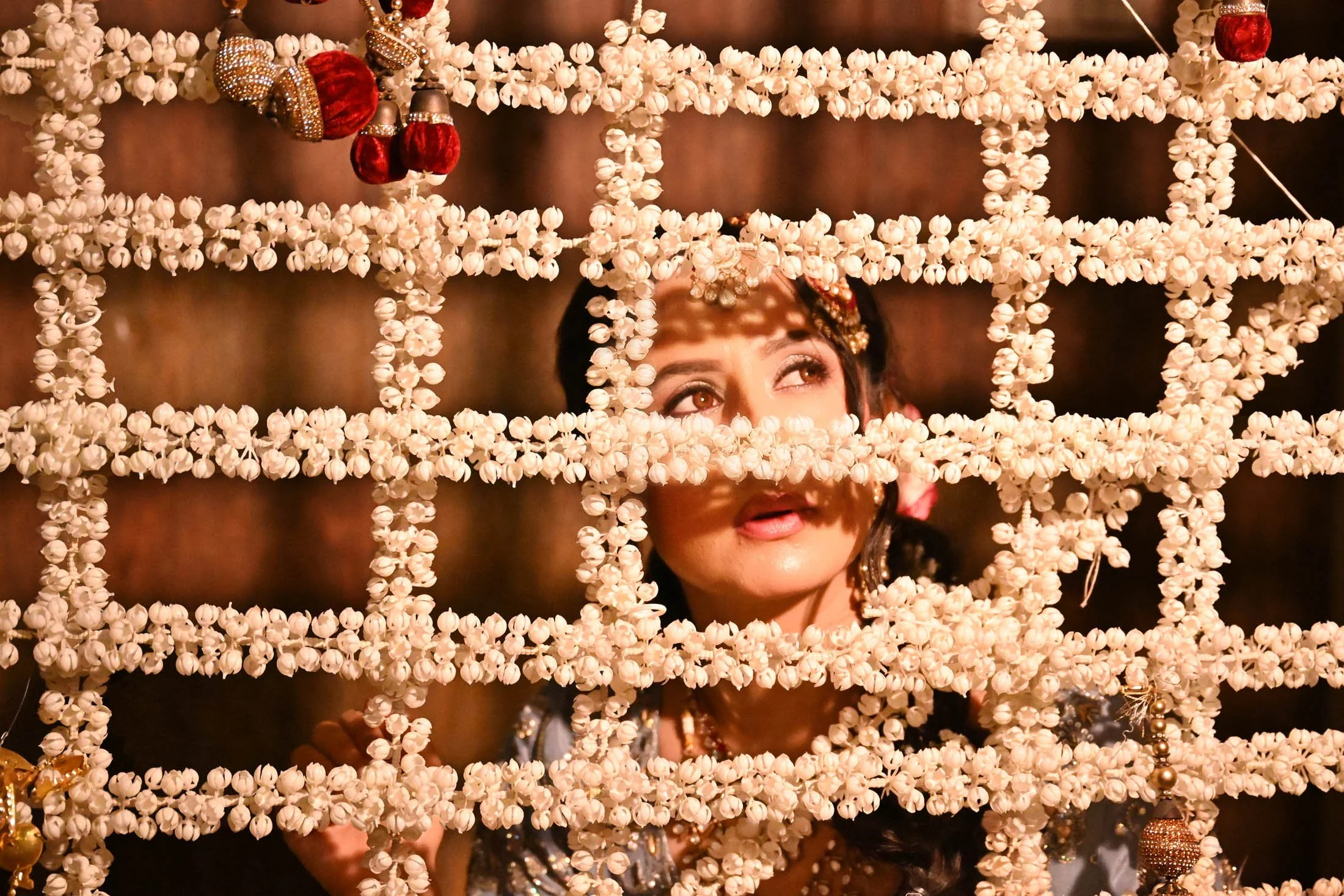 A woman looking through a decorative structure made of small white flowers, with red and gold ornaments hanging from it.
