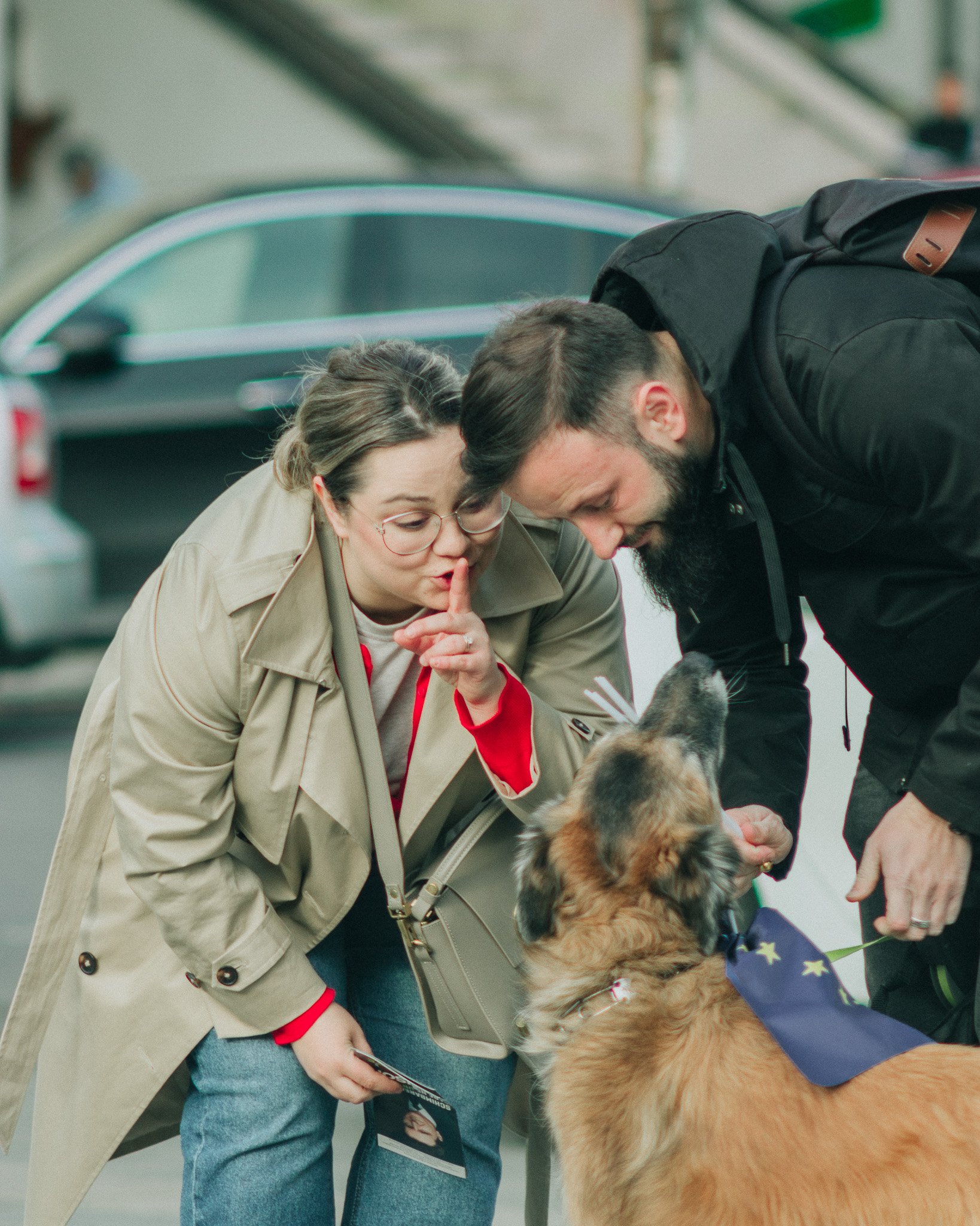 A woman with glasses and a beige trench coat is making a shushing gesture while leaning toward a large dog wearing a European Union flag bandana, as a man with a beard bends down to pet the dog.