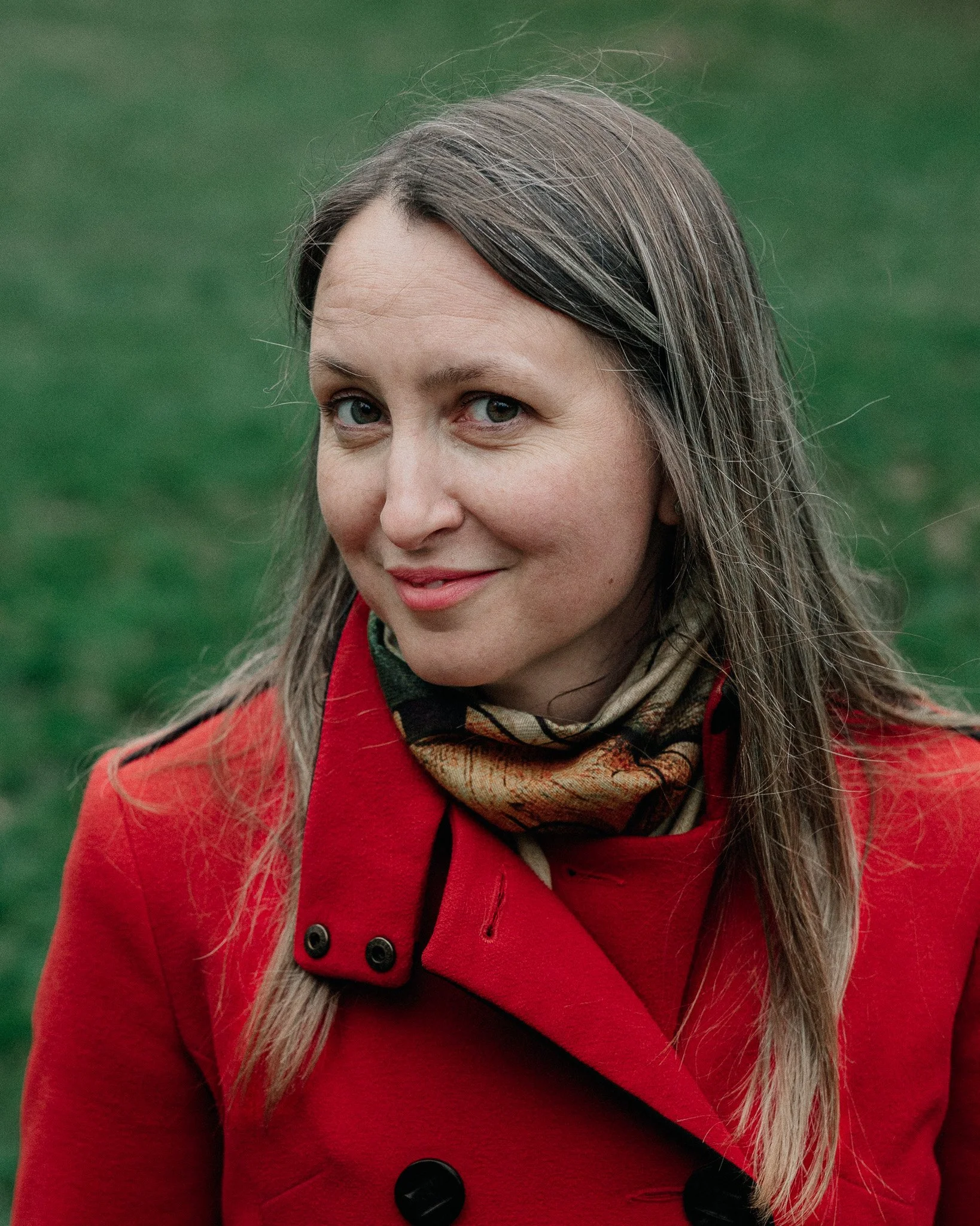 A woman with long light brown hair, wearing a red coat and a patterned scarf, looking at the camera with a slight smile, standing outdoors on green grass.