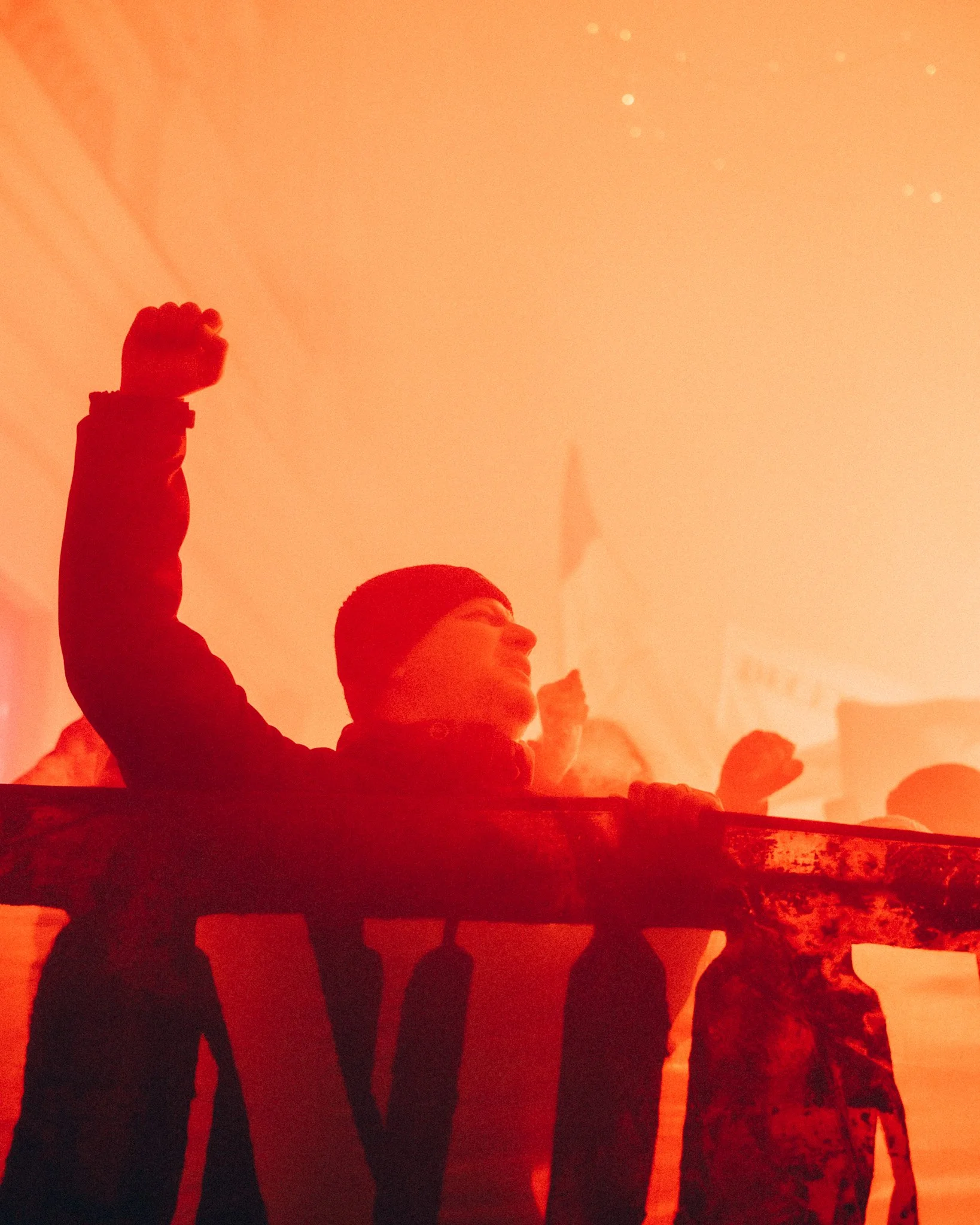 A person with a raised fist standing behind a banner at a protest or rally, with a crowd and flags in the background, colored in red and orange tones.