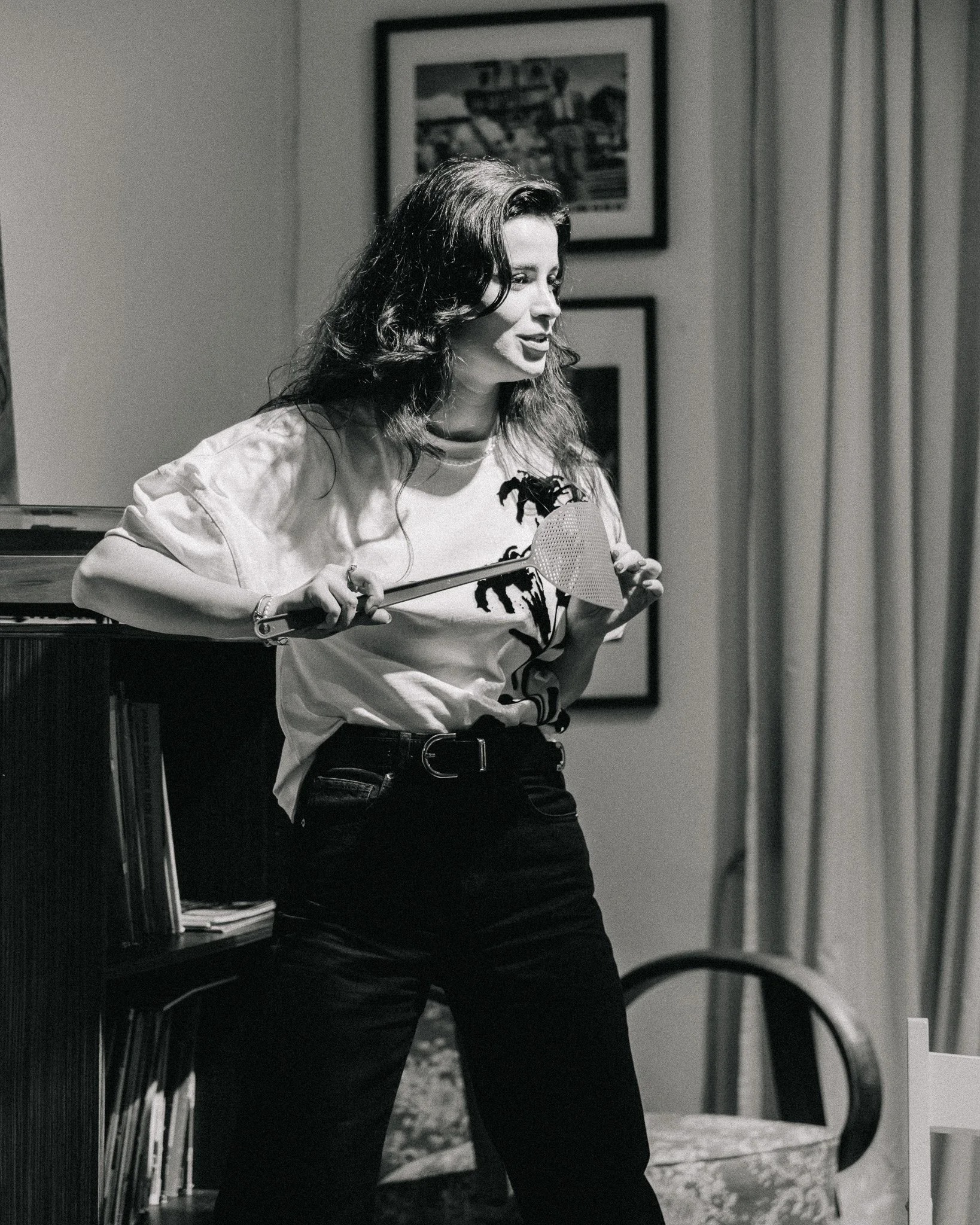 A woman with dark, wavy hair holding a spatula in a home setting, standing near a bookshelf and a chair with a chair cushion, decorated with framed pictures on the wall.