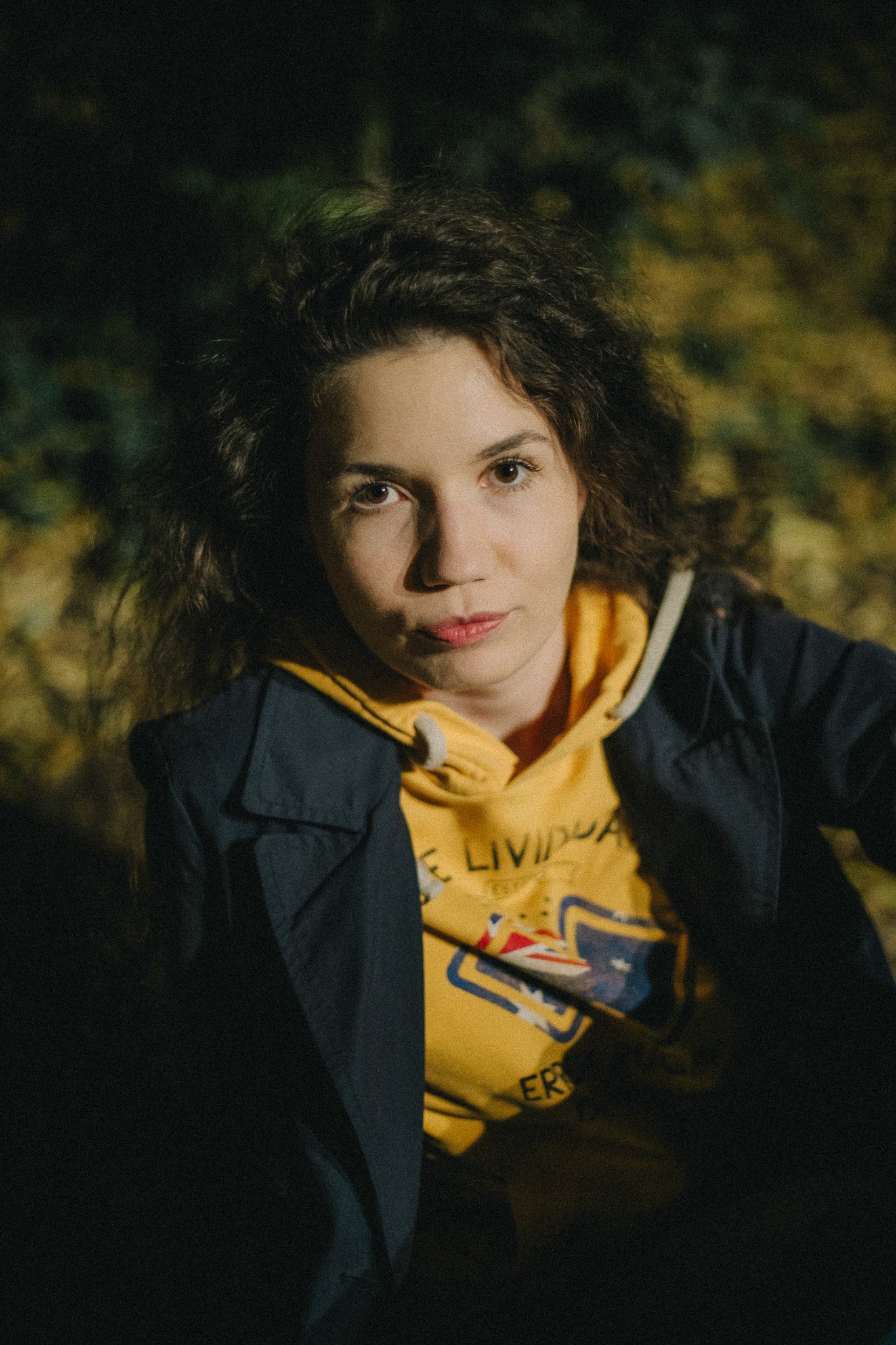A young woman with curly dark hair wearing a yellow hoodie and dark jacket outdoors at night.