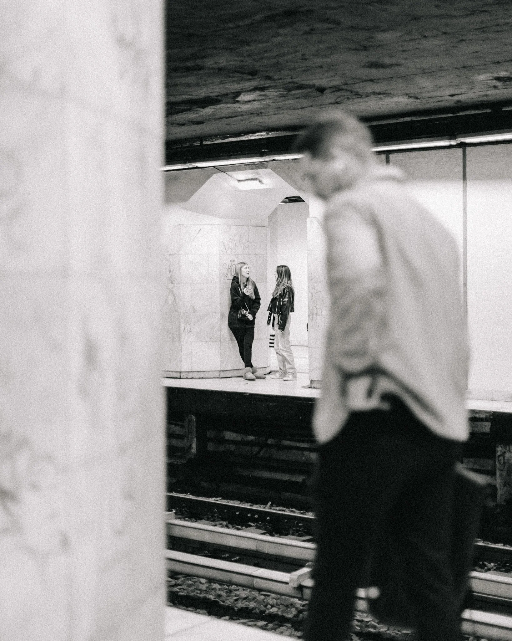 Two women are standing and talking on a subway platform in the background, while a person with glasses is sitting nearby in the foreground.