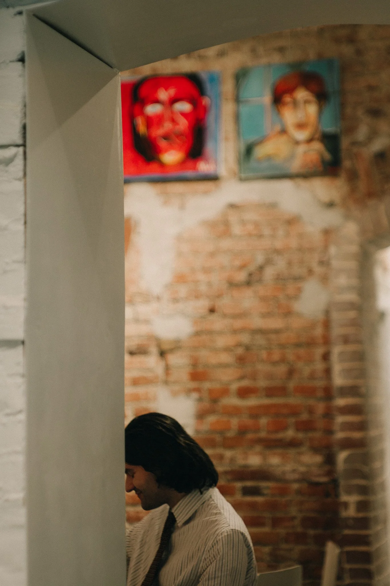 A smiling man with long dark hair, wearing a striped shirt and tie, sitting in a dimly lit room with exposed brick walls and artwork hanging above him.
