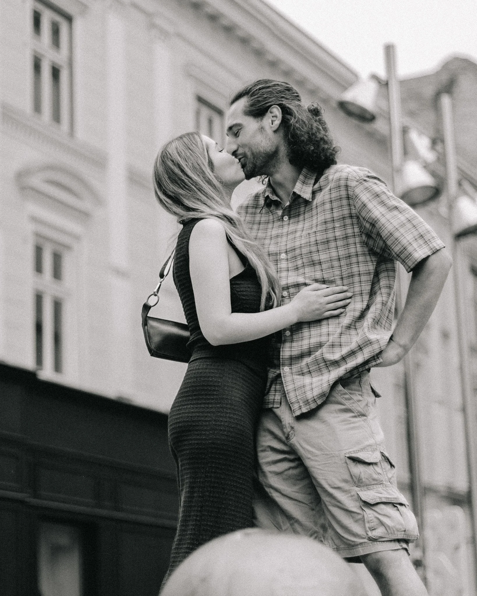 A black-and-white photo of a couple about to kiss on a city sidewalk. The woman is wearing a black sleeveless dress with a shoulder bag, and the man is wearing a short-sleeved plaid shirt and cargo shorts. They are standing close together, with the w