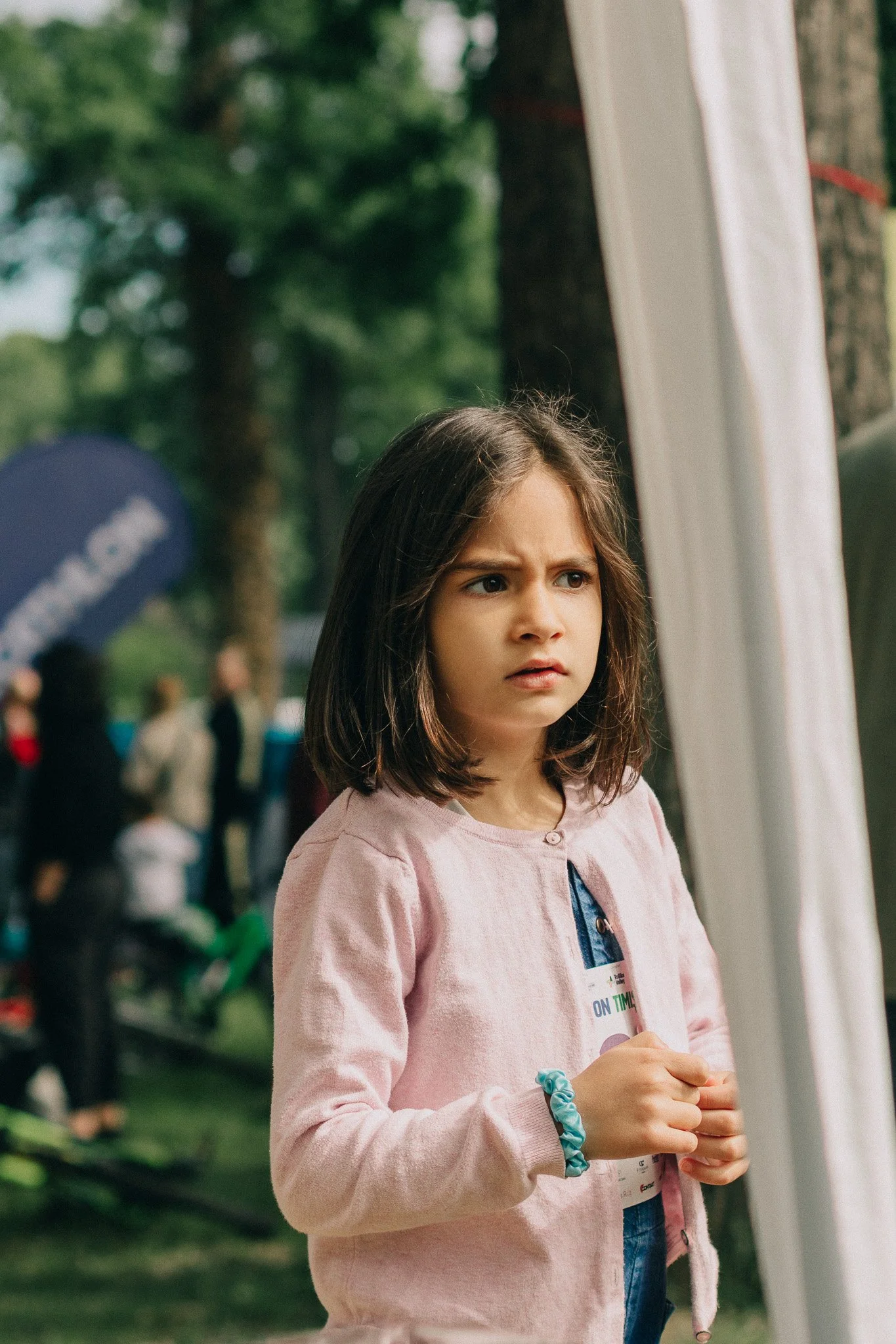 A young girl with shoulder-length brown hair and a pink jacket looks confused or concerned while standing outdoors at an event, with people and trees in the background.