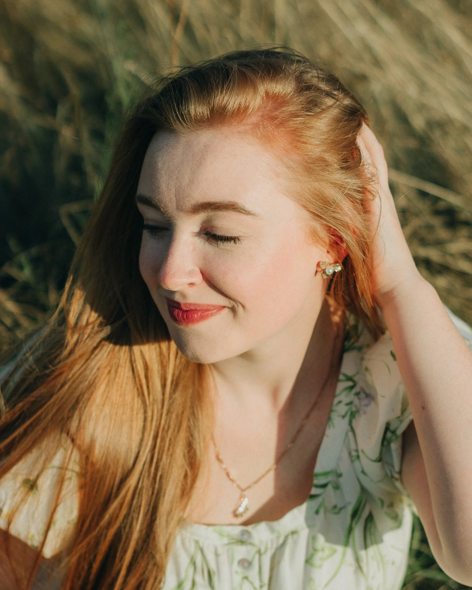 A young woman with red hair, closed eyes, and a gentle smile, sitting outdoors in a field of tall grass, with one hand resting behind her head.