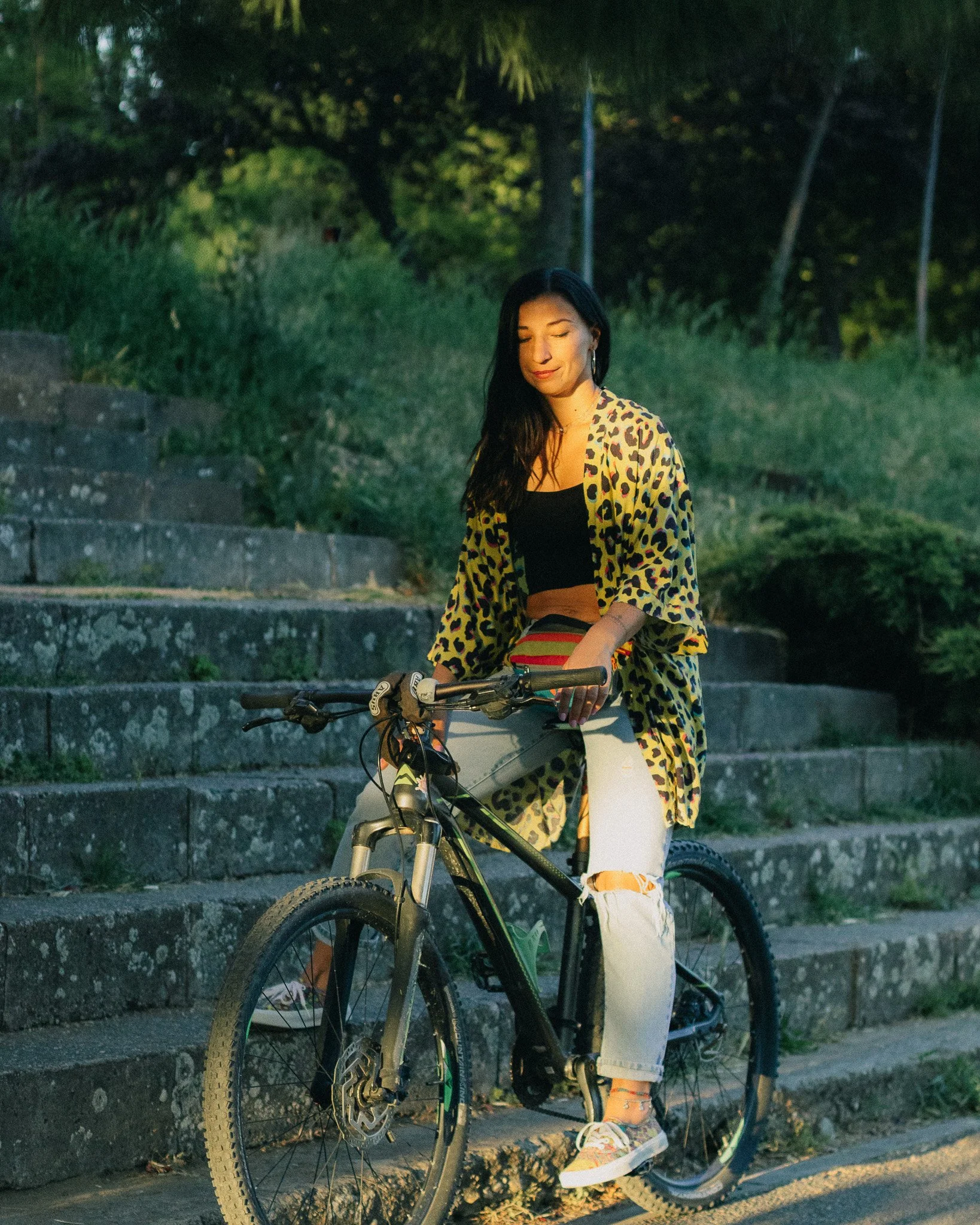 A young woman with long dark hair riding a mountain bike along a stone staircase outdoors during twilight. She is wearing a leopard print jacket, a black crop top, ripped light-wash jeans, and colorful sneakers. The background features green trees an