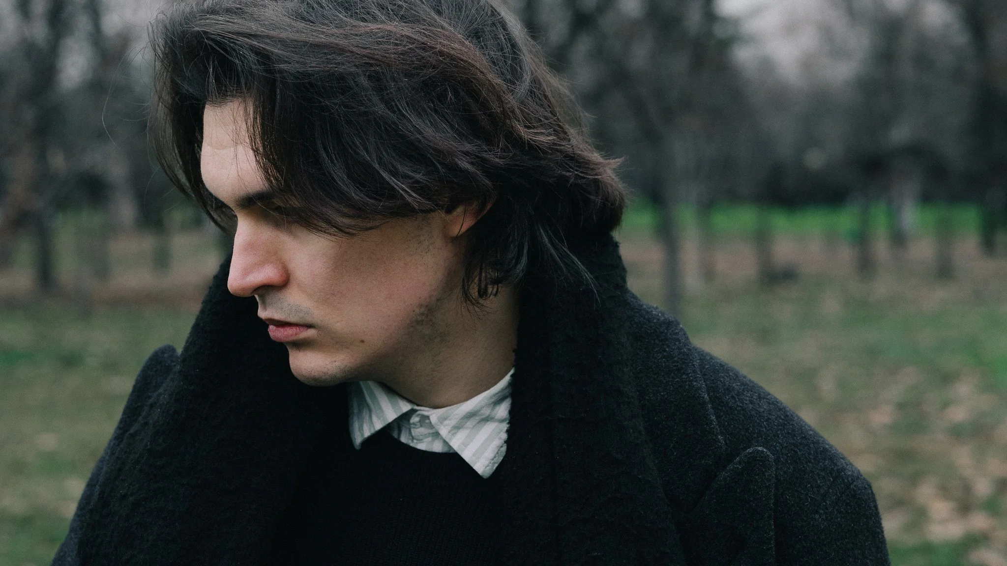 A young man with dark, wavy hair looking down outdoors with trees in the background. He is wearing a dark coat and a striped collared shirt.