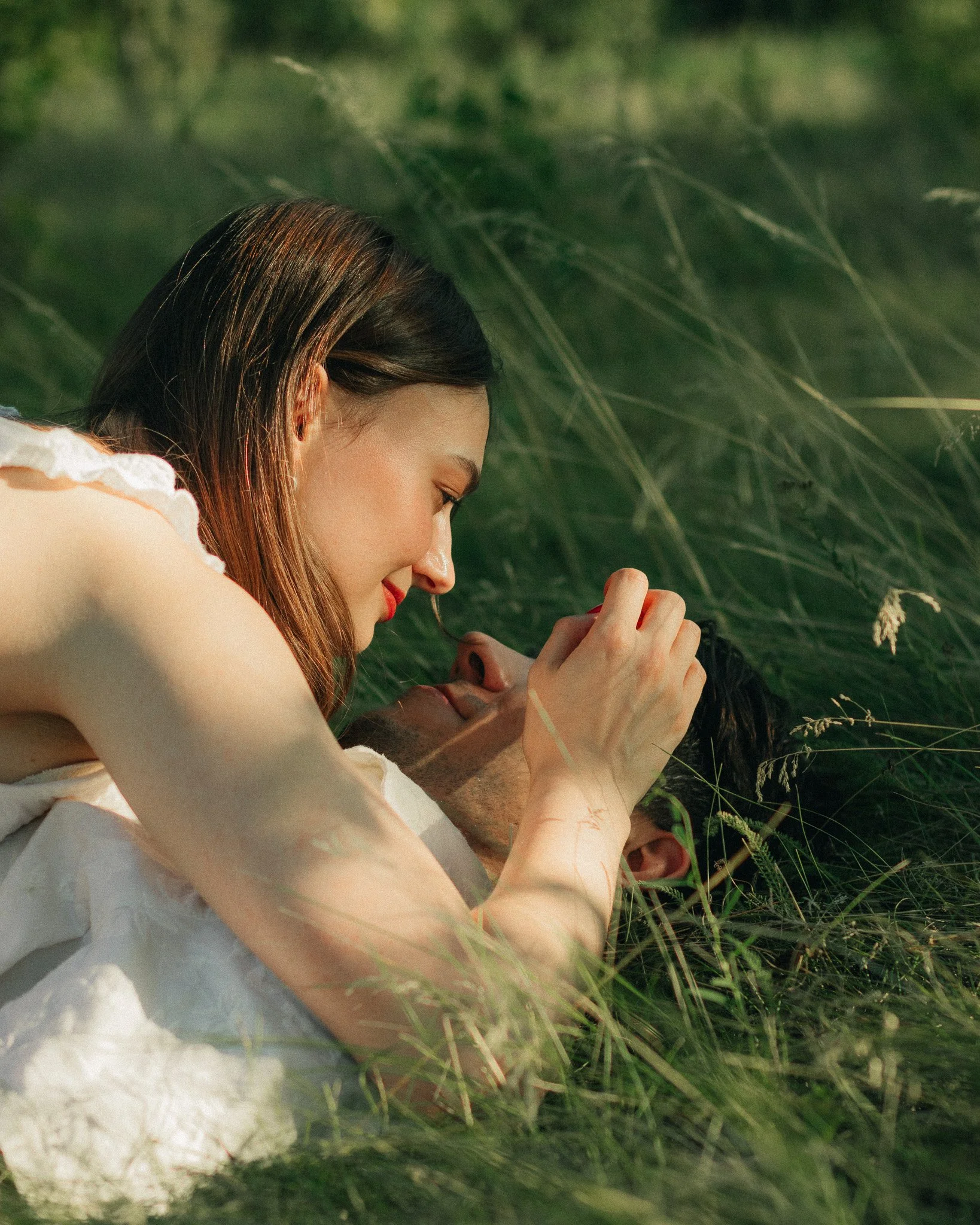 A woman and a man lying in a grassy field, with the woman close to the camera and the man lying on his back, both appearing relaxed and intimate.