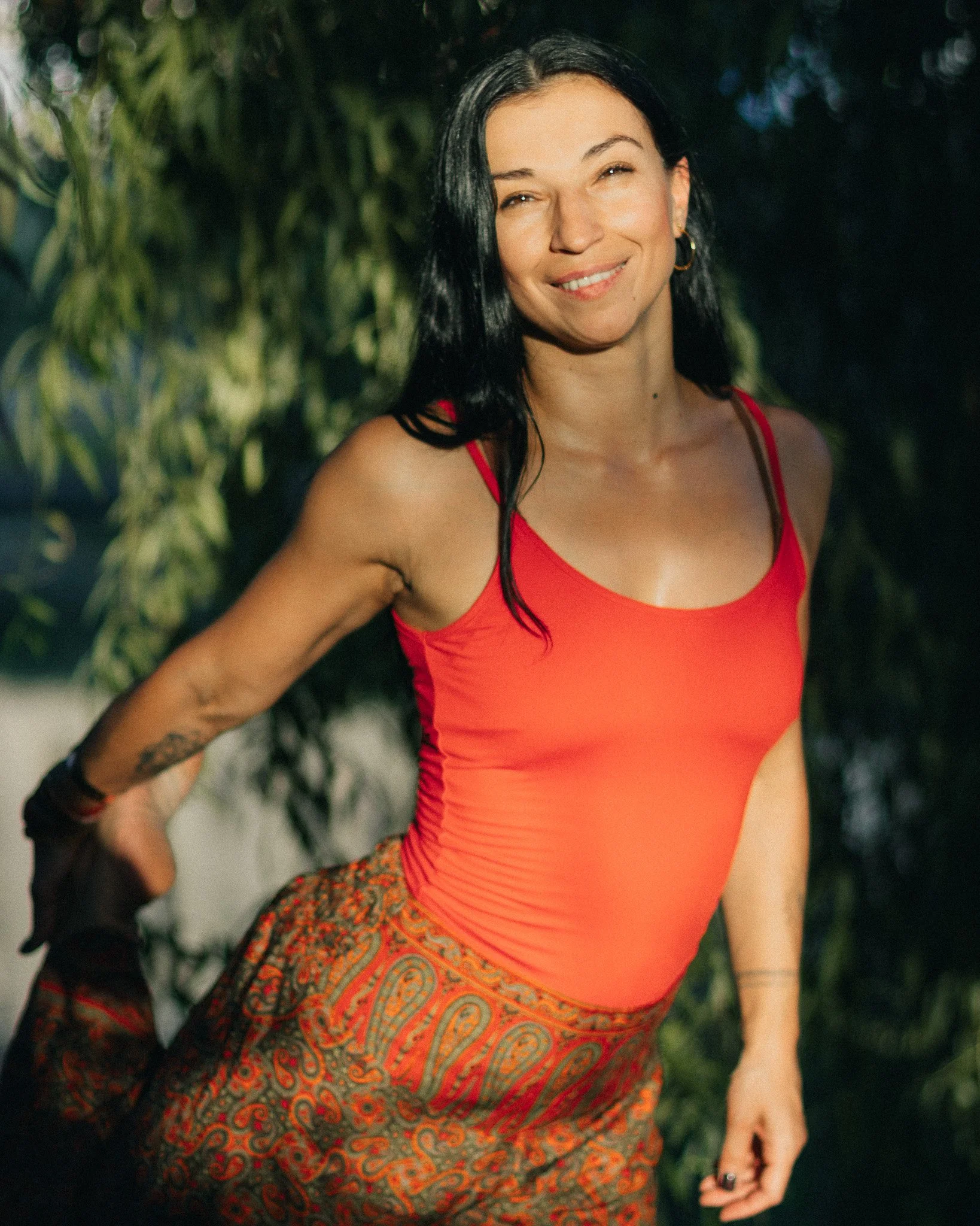 A woman with black hair and light skin, wearing a red tank top and patterned pants, smiling outdoors with green foliage in the background.