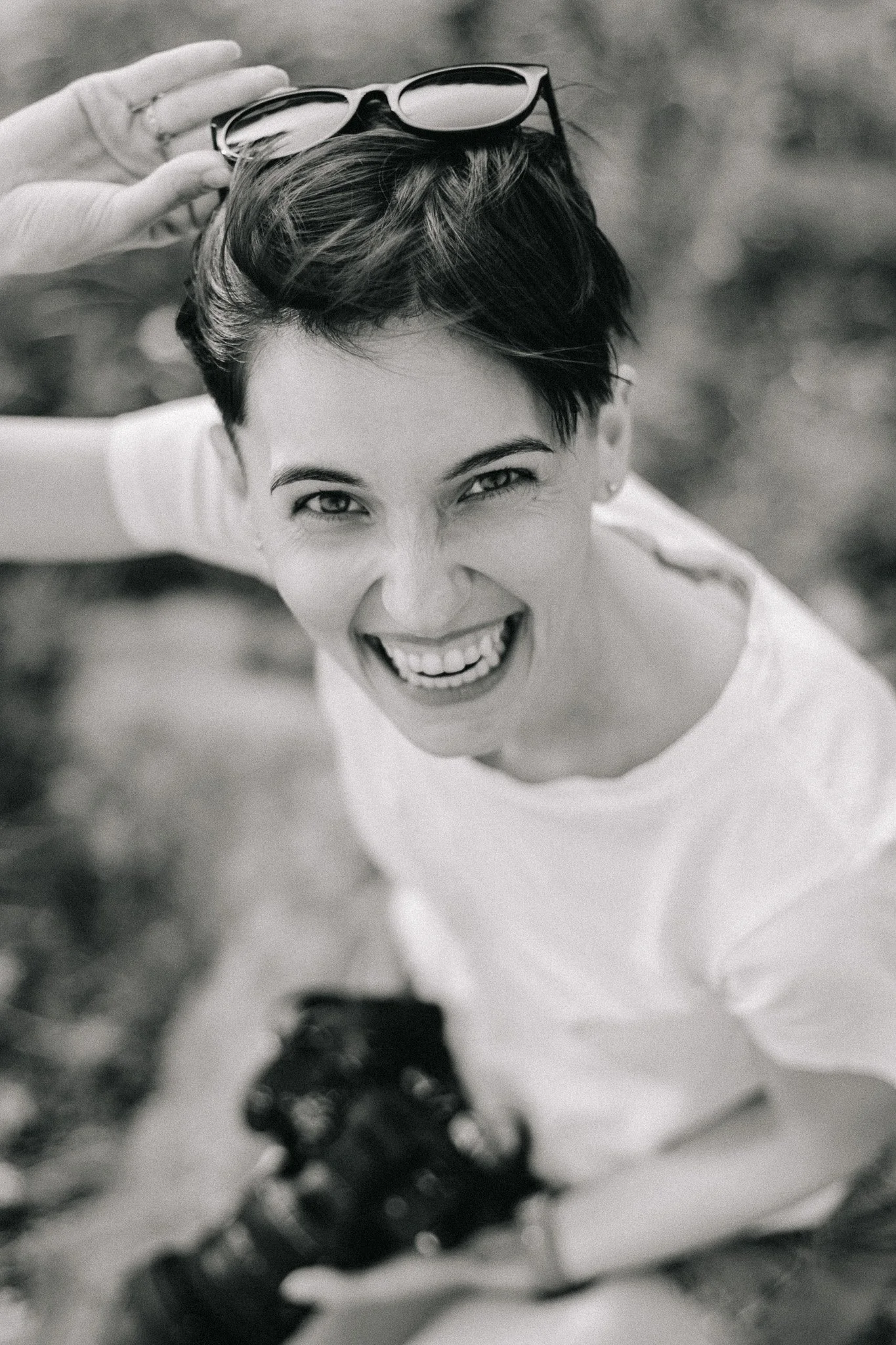 A woman with short hair laughs and looks up at the camera, holding sunglasses on her head, outdoors.