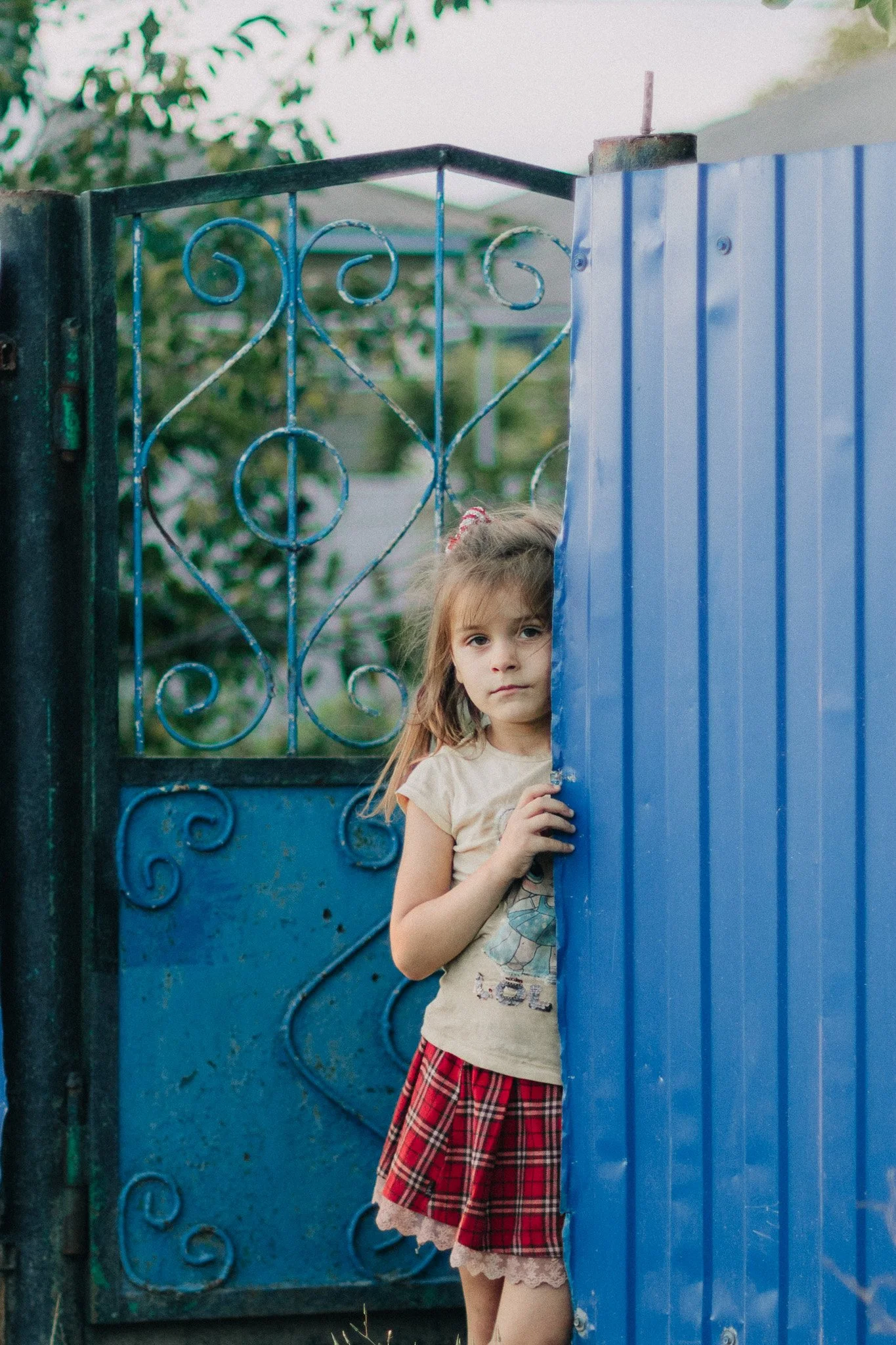 A young girl with long hair and a red plaid skirt peeks from behind a blue metal sheet, standing next to a decorative metal gate, with trees in the background.