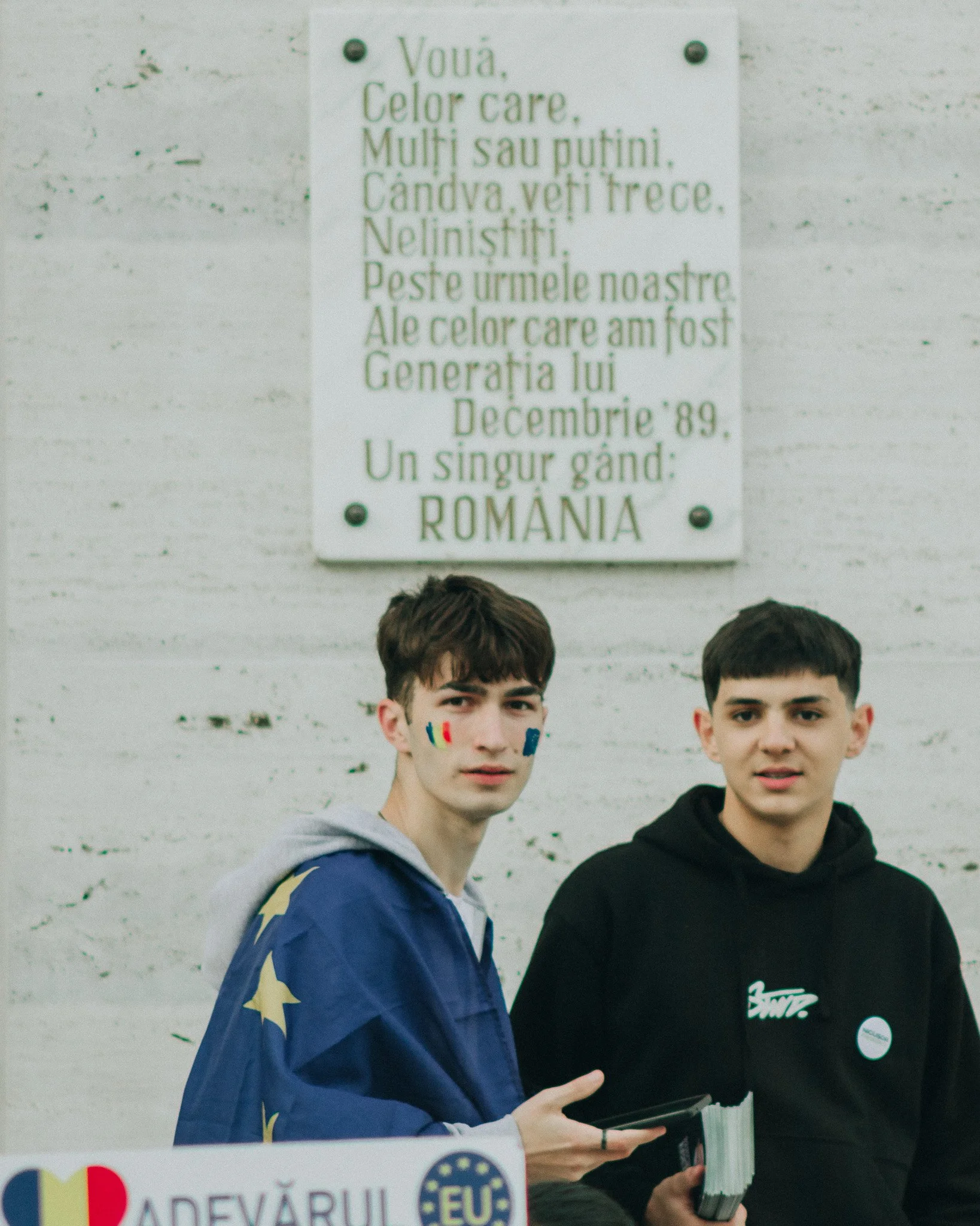 Two young men standing in front of a white wall with a Romanian inscription. One has face paint and wears a flag-themed jacket, the other holds a book or pamphlet.