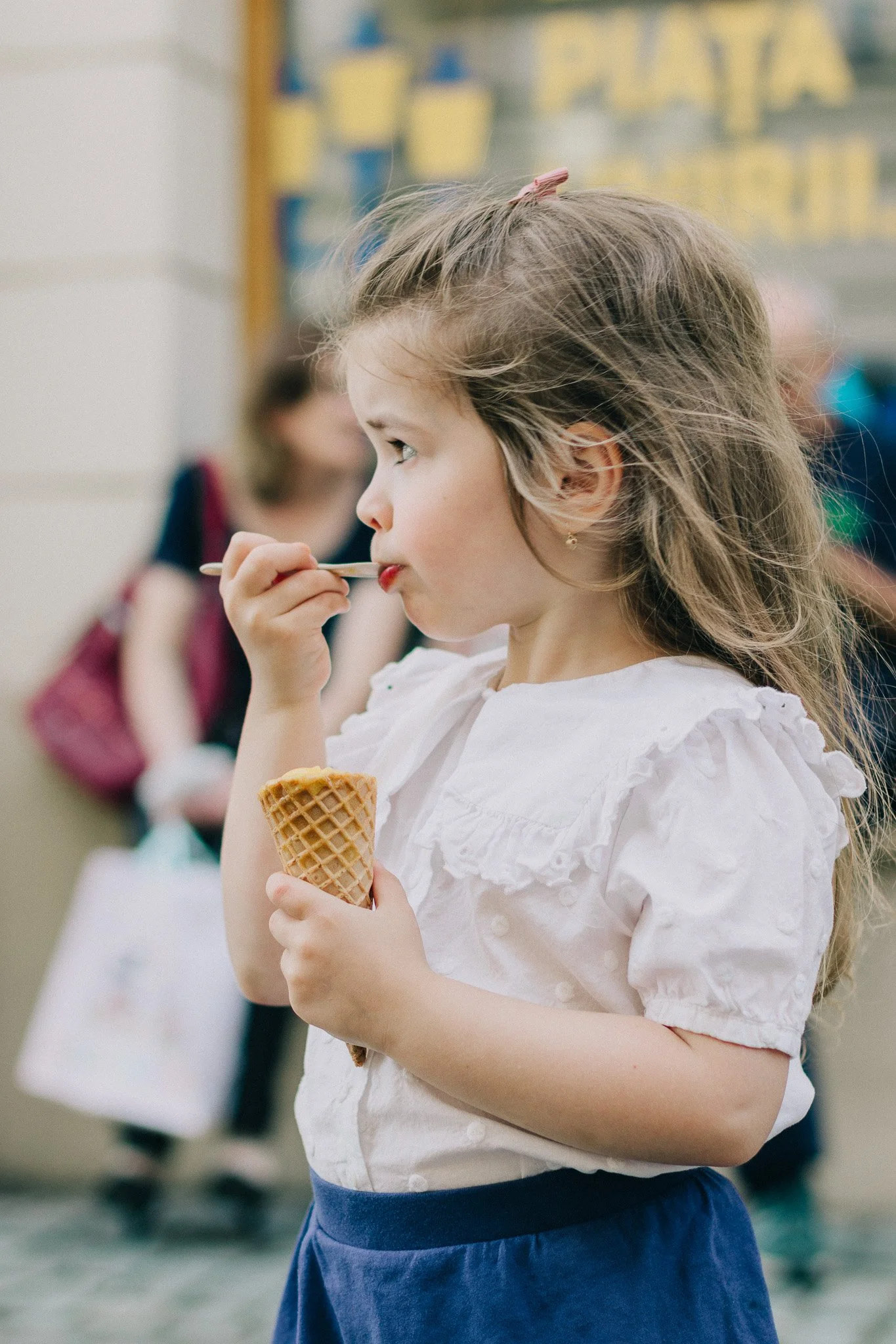 A young girl with long, wavy hair, wearing a white blouse and blue skirt, eating ice cream cone in a busy indoor setting.