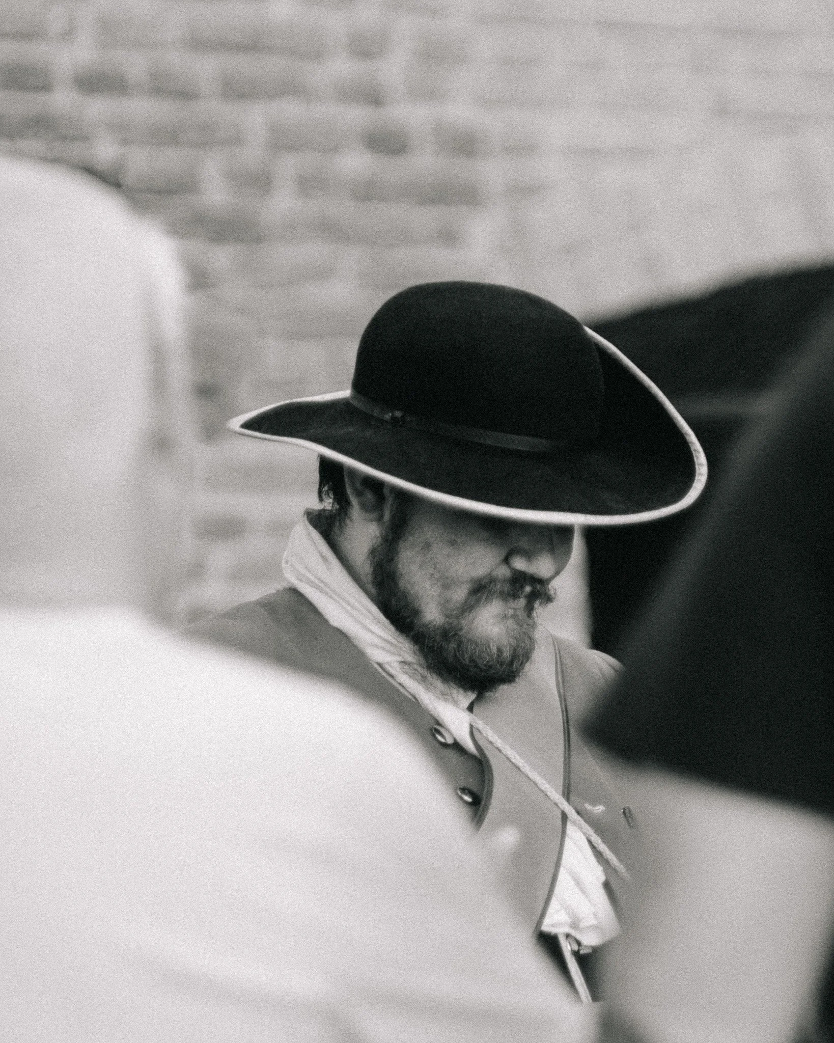 Man wearing a large black hat with a wide brim, beard, and mustache, looking downward, with blurred people in the foreground and a brick wall in the background.