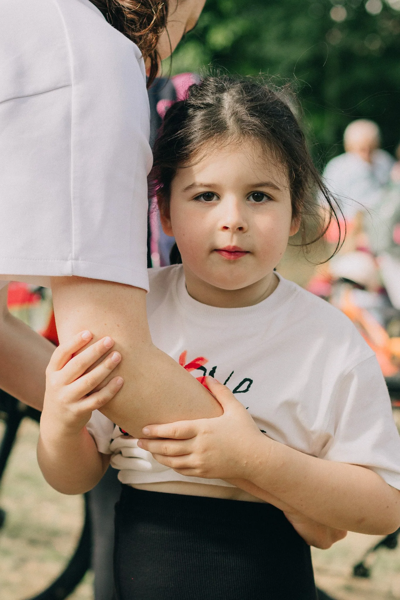 A young girl with brown hair, wearing a white T-shirt, is holding her arm and looking at the camera. An adult woman, partially visible, is holding her arm and supportive. Background shows a blurred outdoor setting with people and greenery.