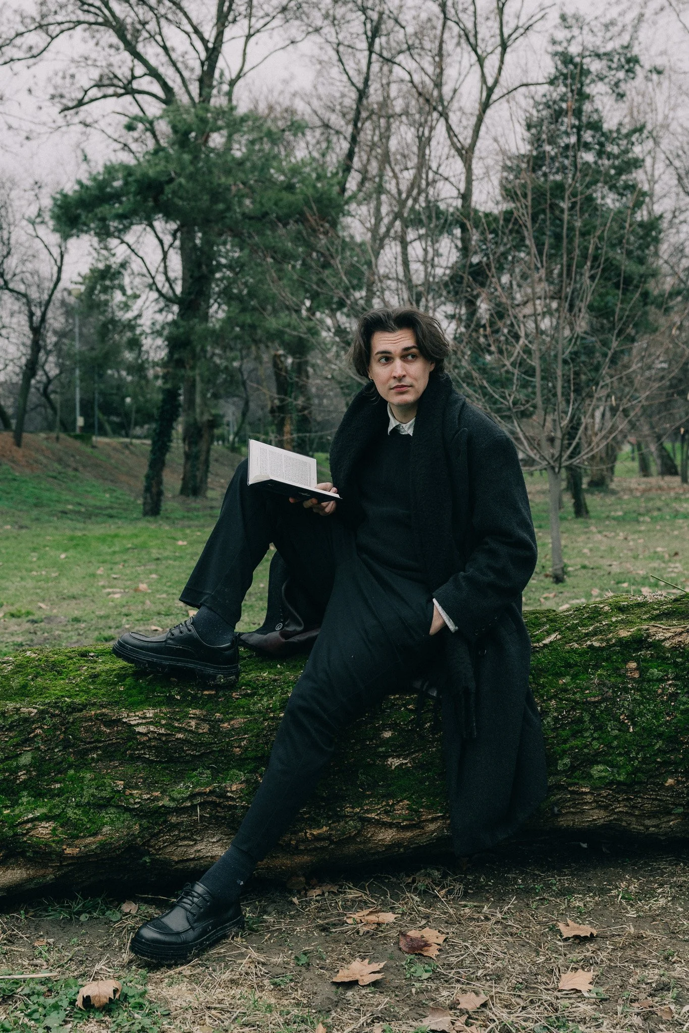 A young man with dark wavy hair, wearing black clothing and shoes, sitting on a large moss-covered log in a park, holding a book, with trees and overcast sky in the background.