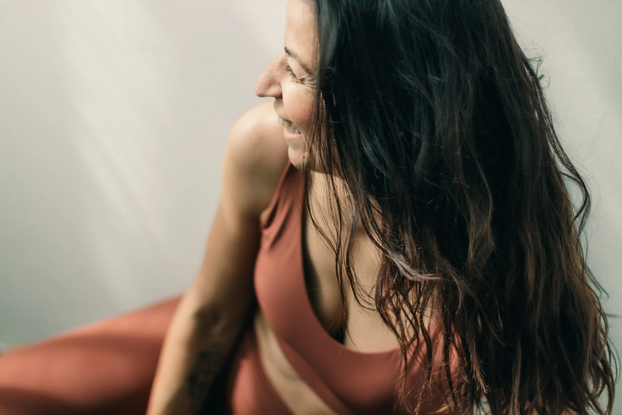 A woman with long, wavy dark hair wearing a sleeveless, rust-colored top, smiling and looking down.