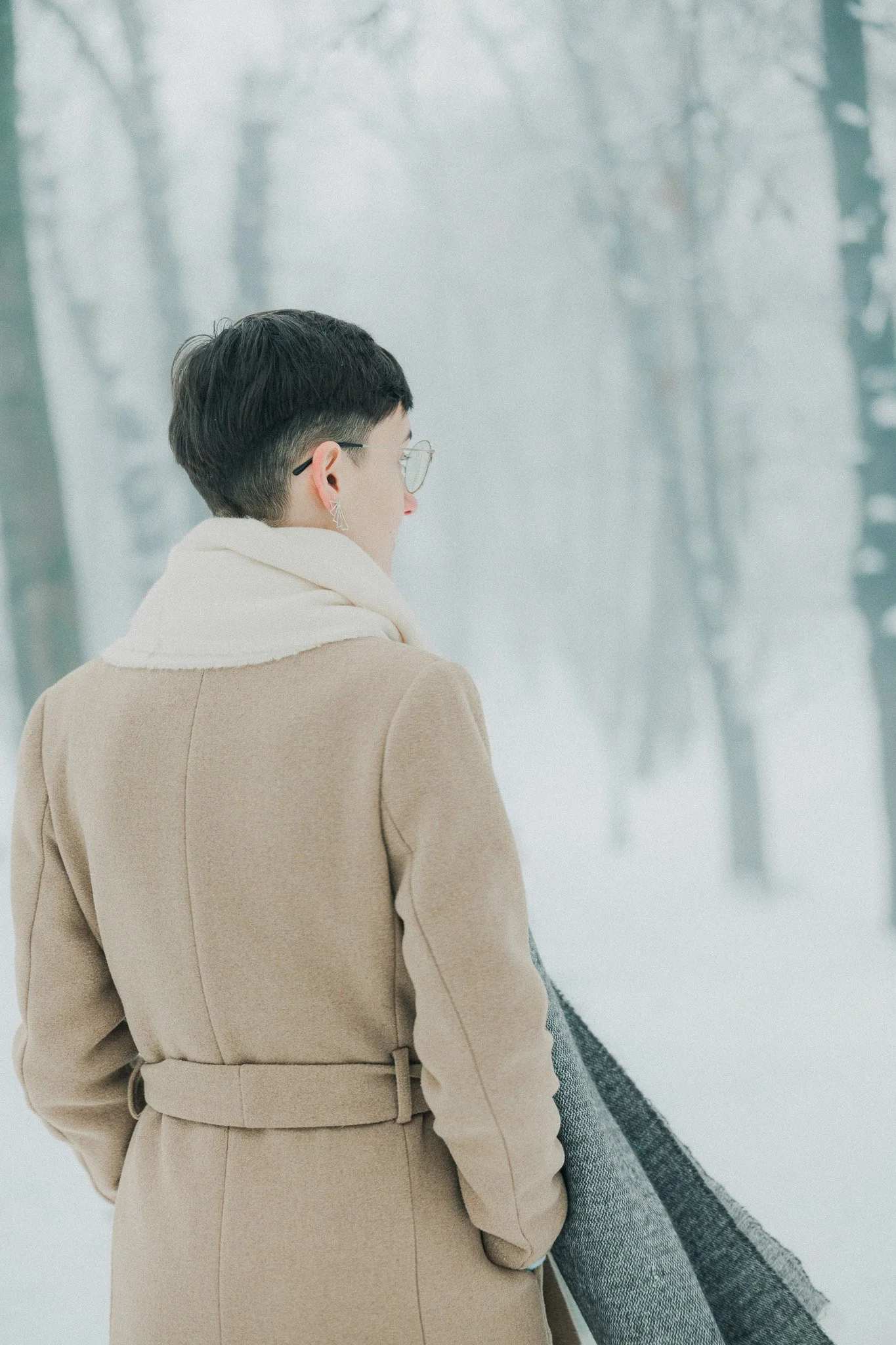 A woman with short dark hair, glasses, and star earrings, wearing a beige coat and white scarf, standing in a snowy forest.