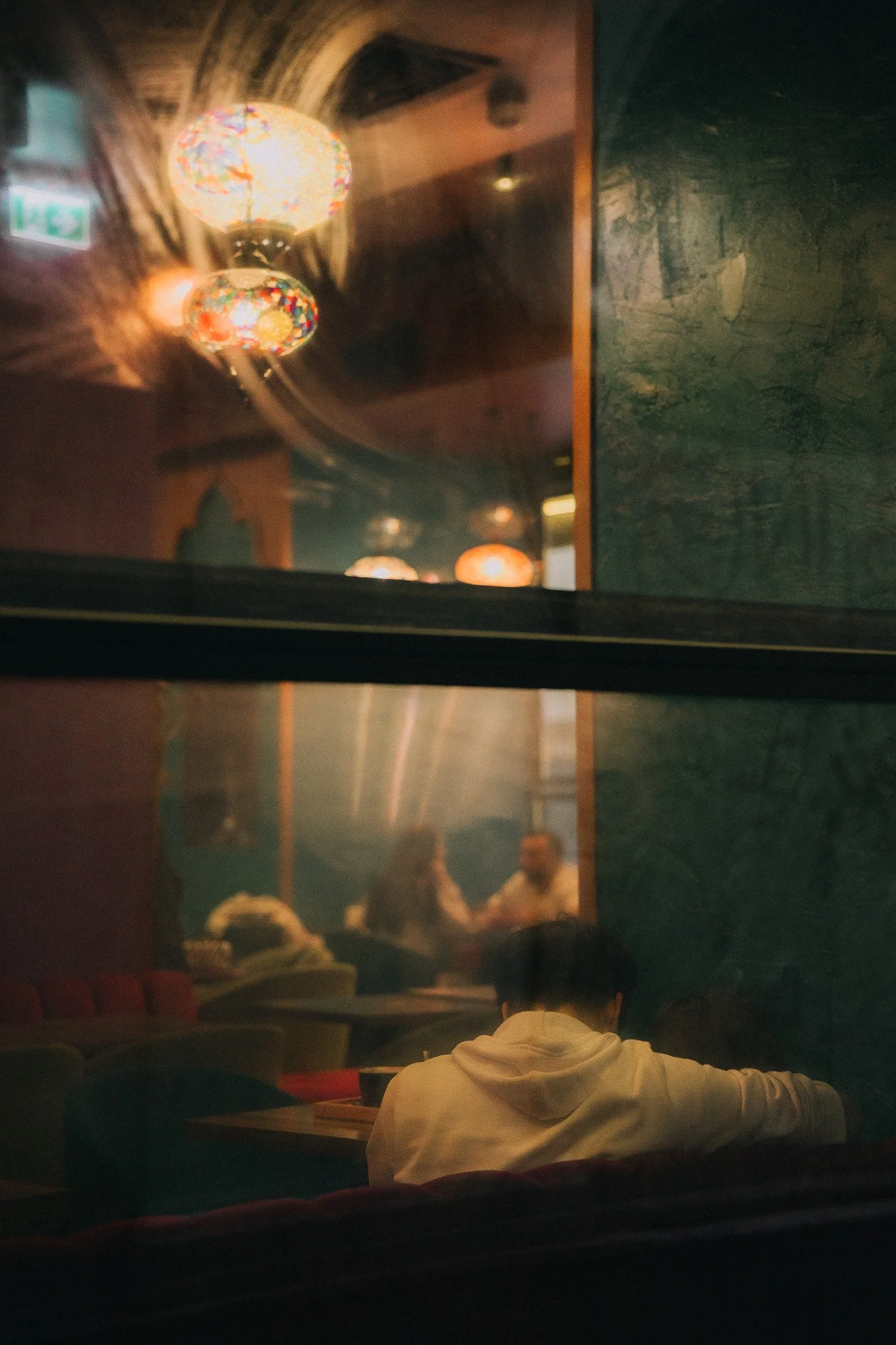 A reflection of colorful hanging lamps on a glass window reveals people sitting inside a dimly lit restaurant or café, with some individuals engaged in conversation and one person wearing a white hoodie sitting at a table.