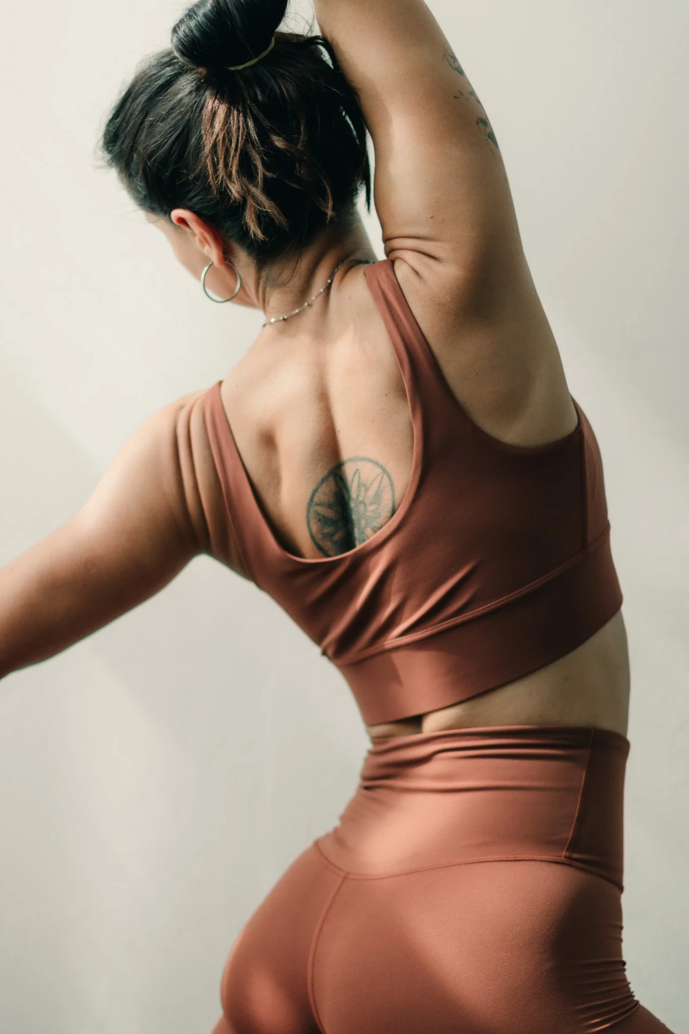 A woman with short dark hair, tattoos, and hoop earrings, wearing a brown athletic top and matching high-waisted leggings, is posing with her right arm raised above her head.