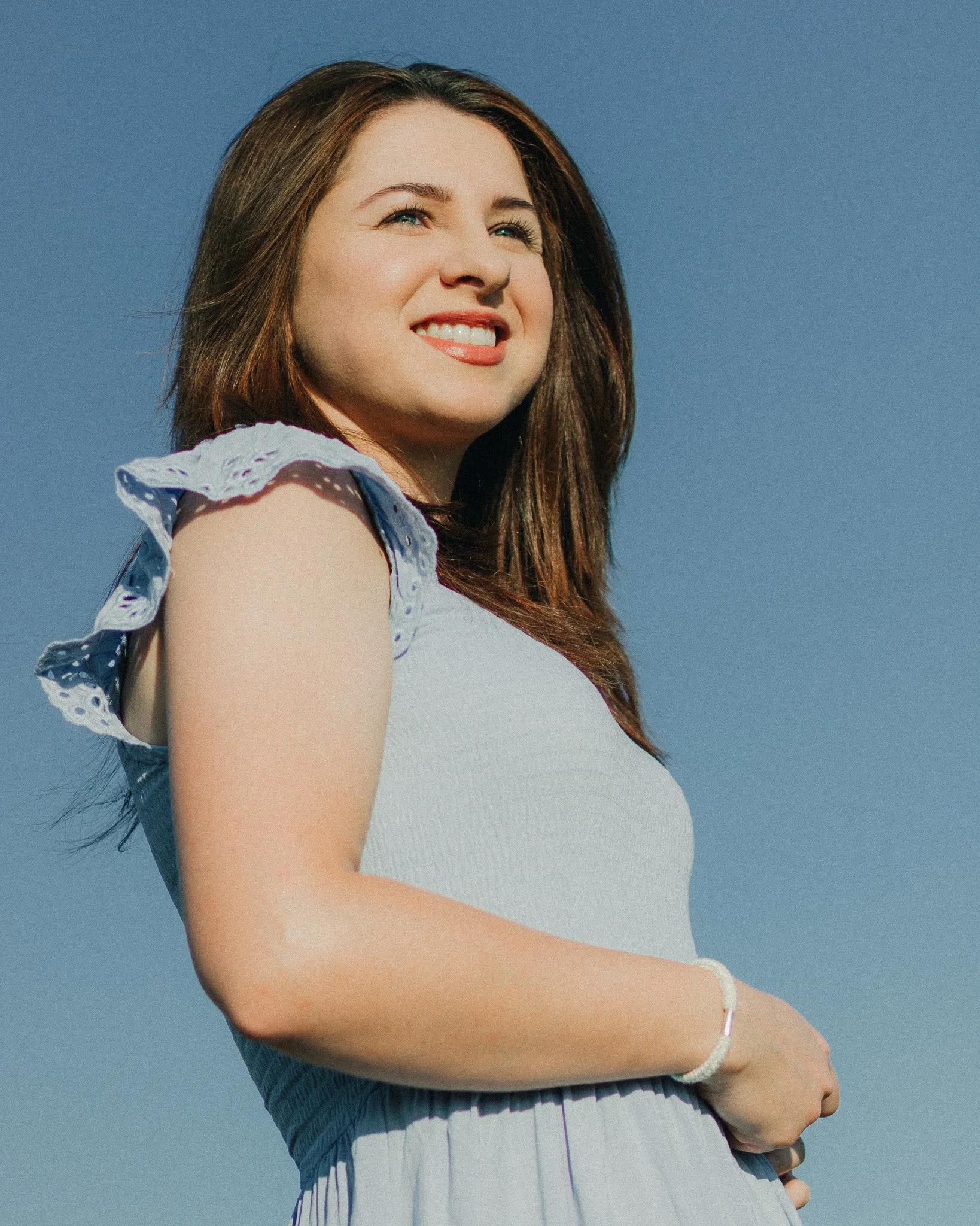 A smiling woman with brown hair, wearing a light-colored dress with ruffled sleeves, standing outdoors against a clear blue sky.