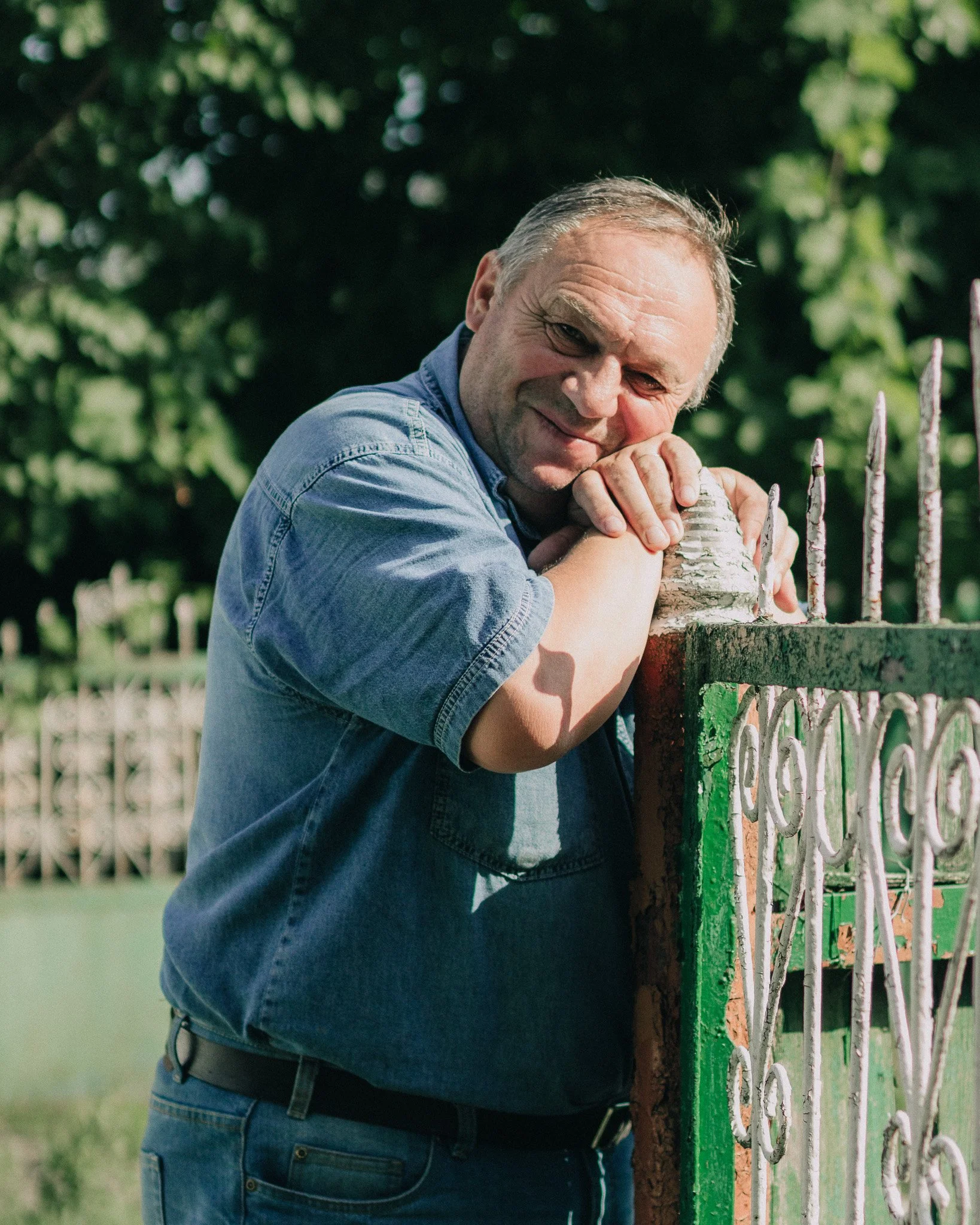 A middle-aged man leaning on a decorative metal fence, smiling and looking at the camera, with green trees in the background.