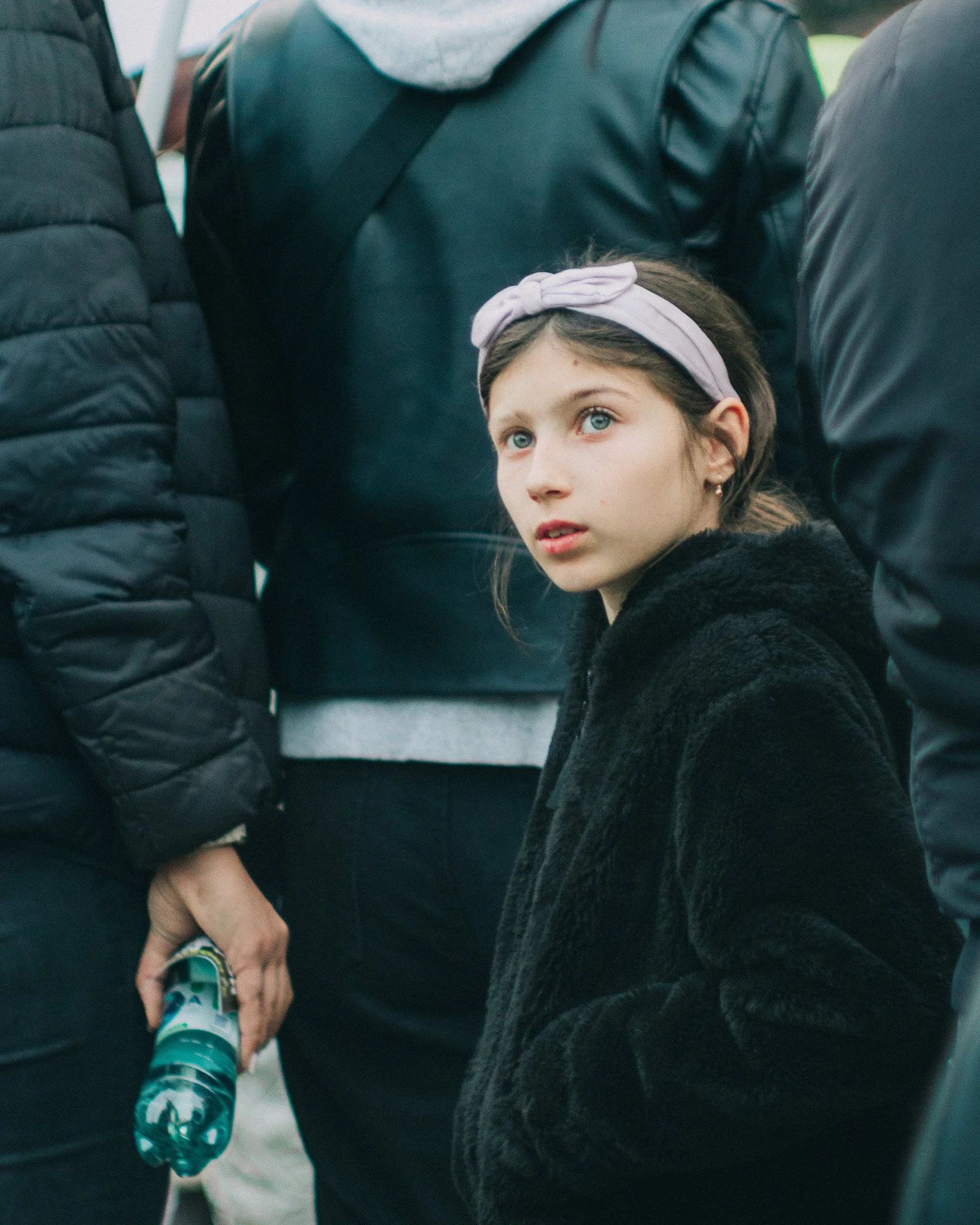 A young girl with blue eyes, wearing a light purple headband and a black fleece jacket, looks to the side among a group of people.